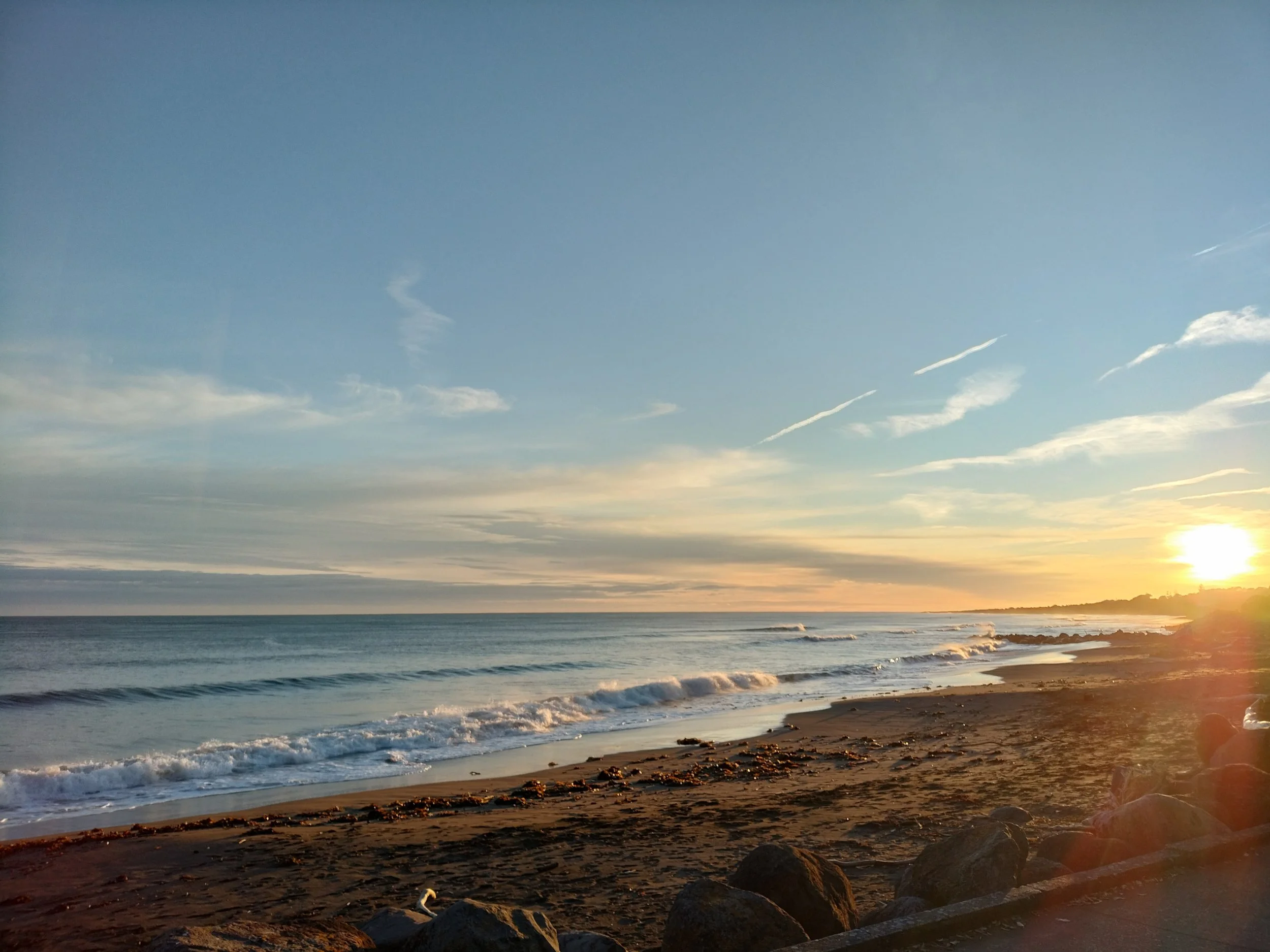 A sunset over the ocean with the sun low on the horizon, sending warm golden light over the water and shoreline. The sky is mostly clear with some wispy clouds.