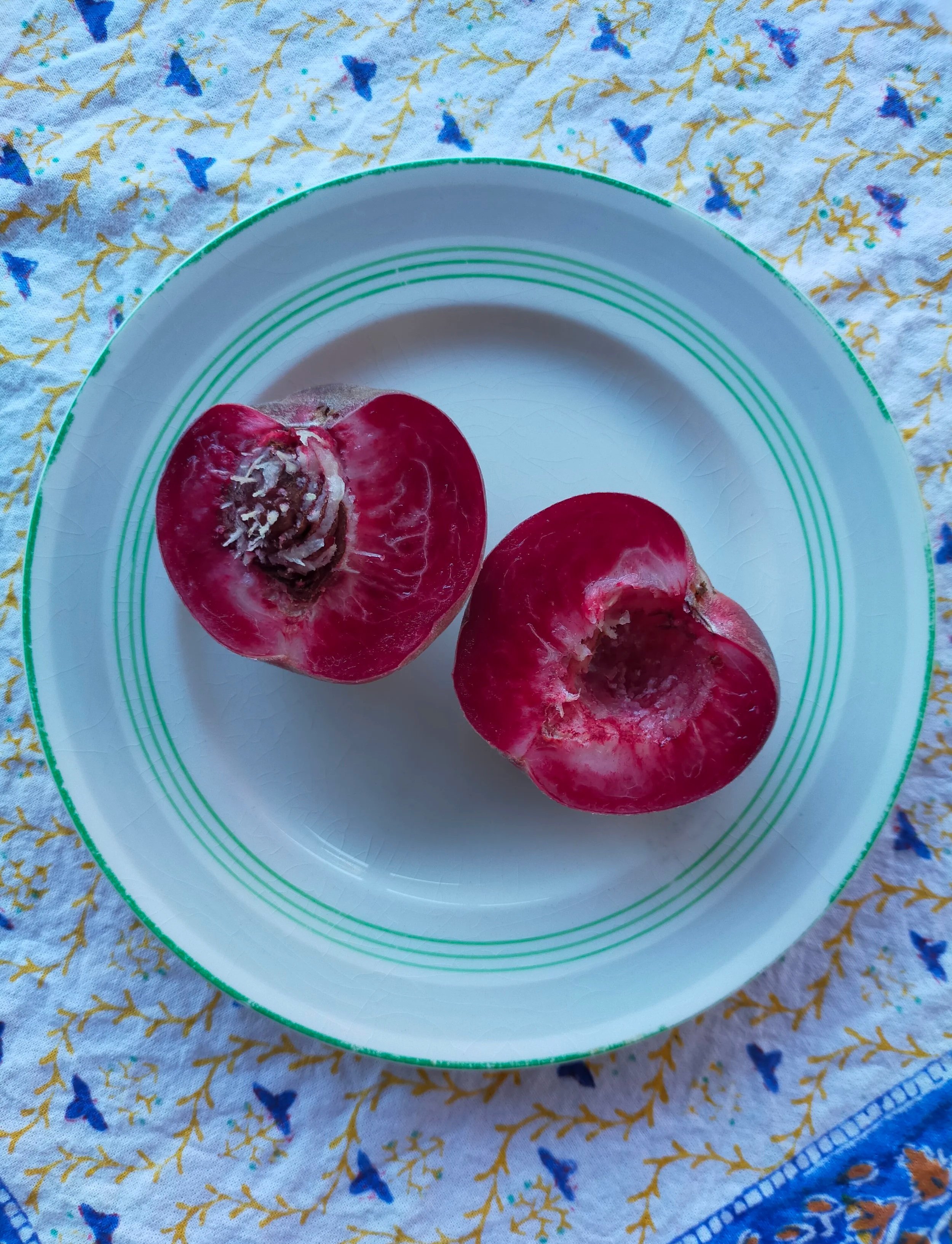 A white plate with green stripes contains two pomegranate halves on a tablecloth decorated with small blue birds and yellow twig patterns.