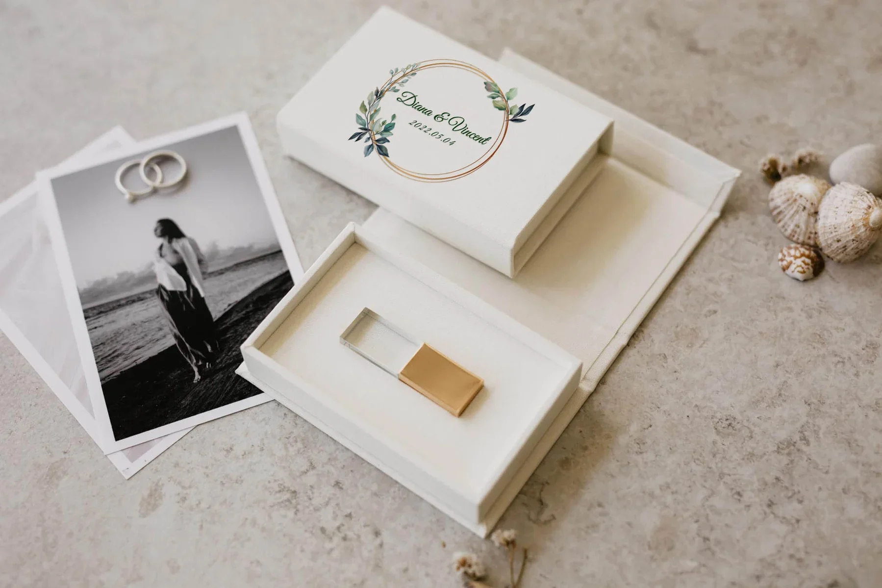 Une boîte blanche avec un design floral contenant des éléments pour un mariage, incluant des photos en noir et blanc d'une femme et deux alliances, ainsi qu'un petit objet rectangulaire en or et transparent. Sur la boîte, il est écrit "Diana & Vincent 2022.05.01".