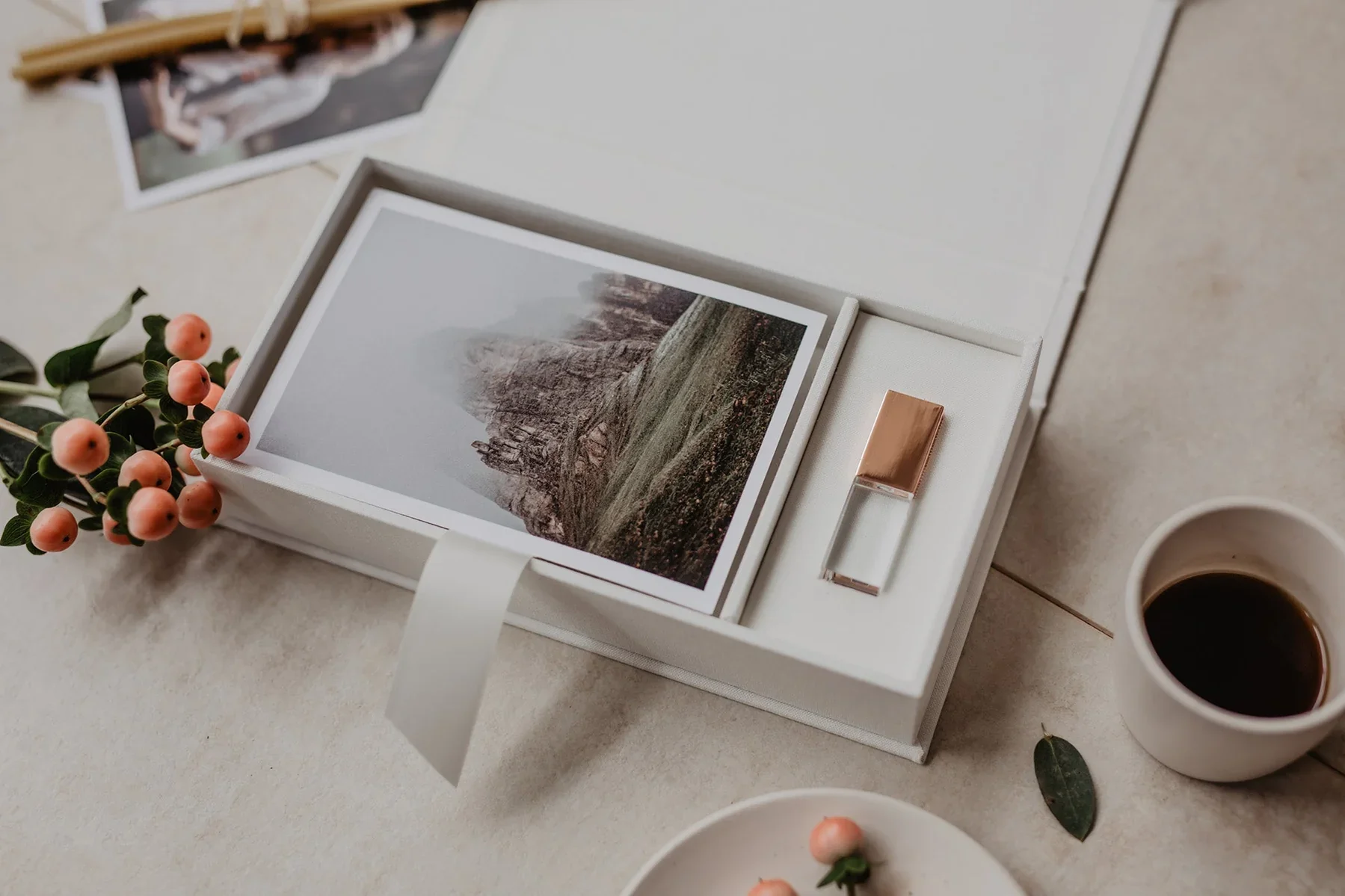 Une boîte blanche avec une photo d'un paysage rocheux, un petit vase avec des baies roses et des feuilles vertes, une tasse de café noir.