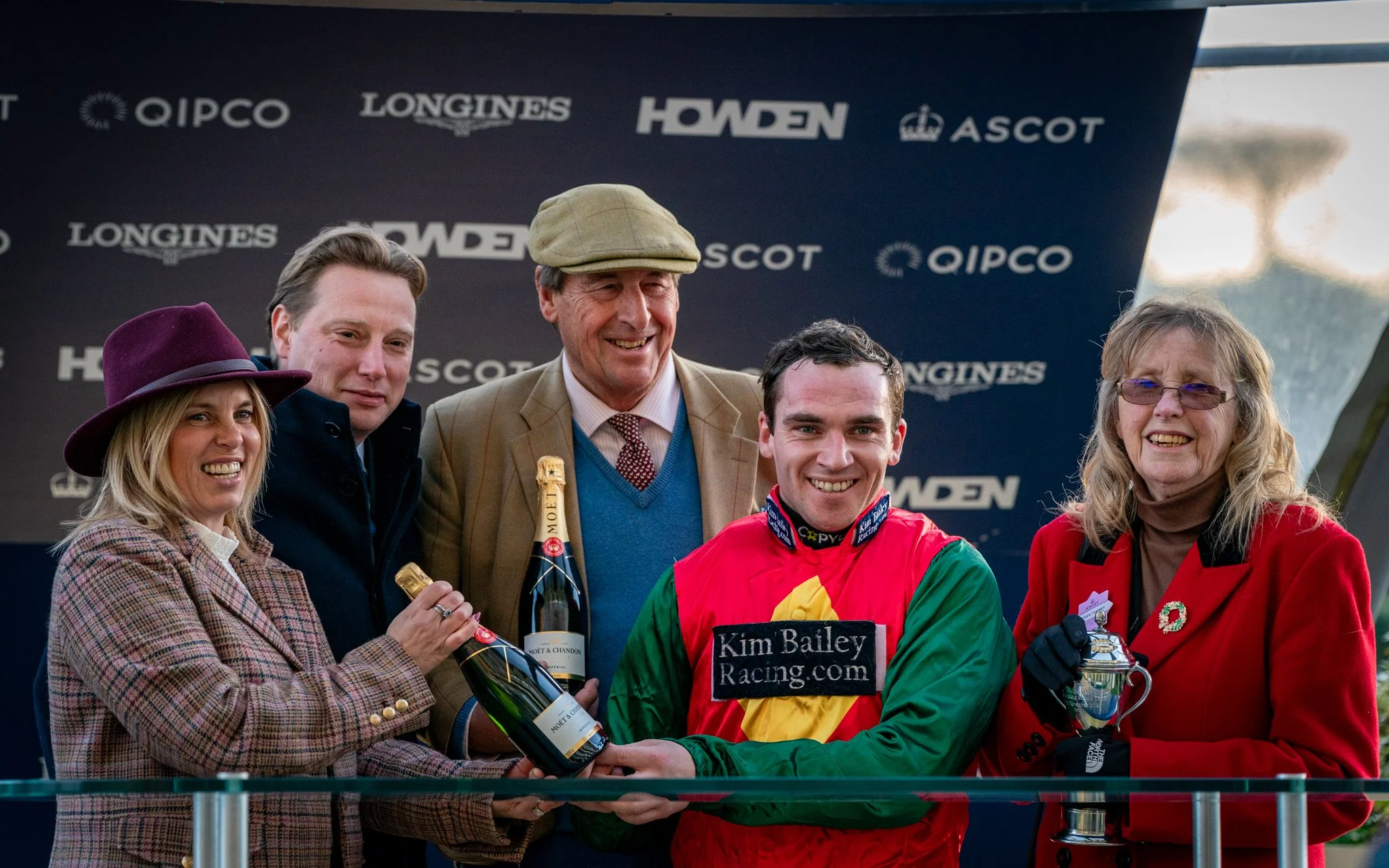 A group of six people, including a jockey wearing colorful racing silks, standing together on a podium with a dark backdrop featuring sponsor logos like QIPCO, Longines, Howden, and Ascot, celebrating a race event.