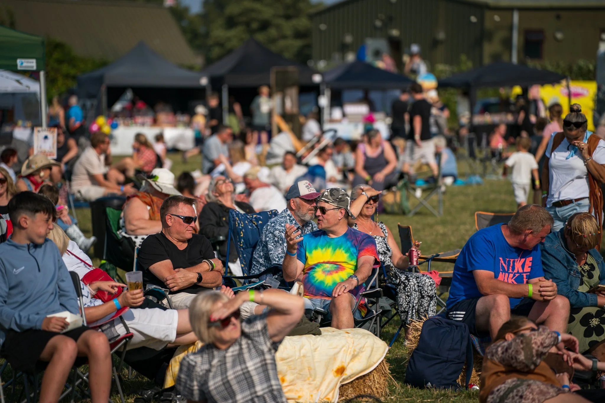 Crowd of people sitting on chairs outdoors during daytime at a festival or event, with tents and green buildings in the background.