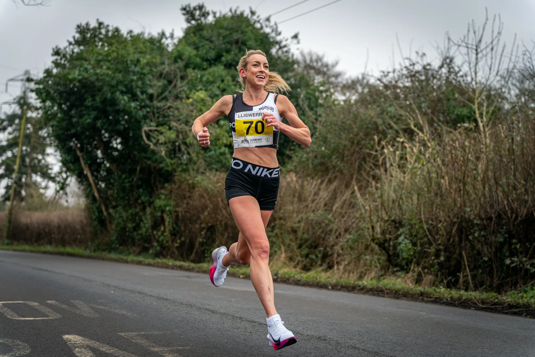 A woman with a race bib numbered 70 running on a road during daytime, wearing a black and white sports top and shorts, with trees and bushes in the background.