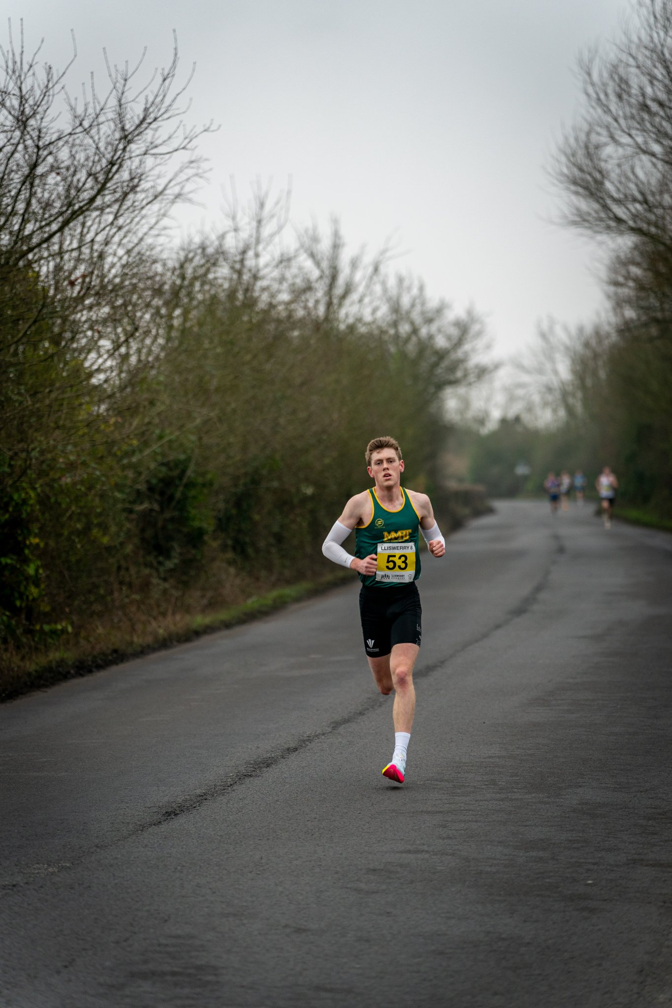 A young male runner in a green and black athletic outfit running on a paved road with trees and other runners in the background.