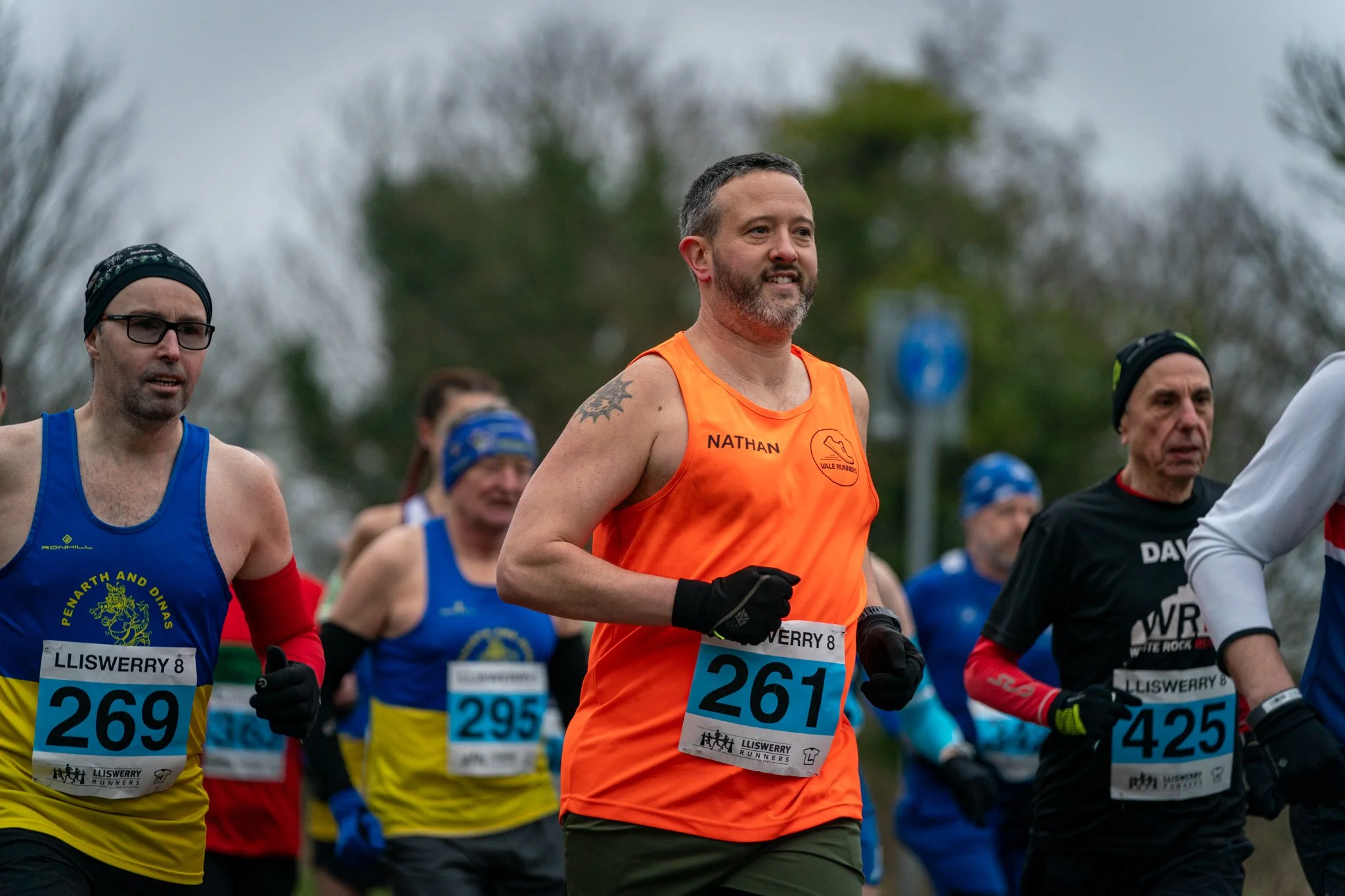 Group of marathon runners participating in a race outdoors, wearing athletic gear with bib numbers, some with hats and gloves, running on a cloudy day with trees in the background.