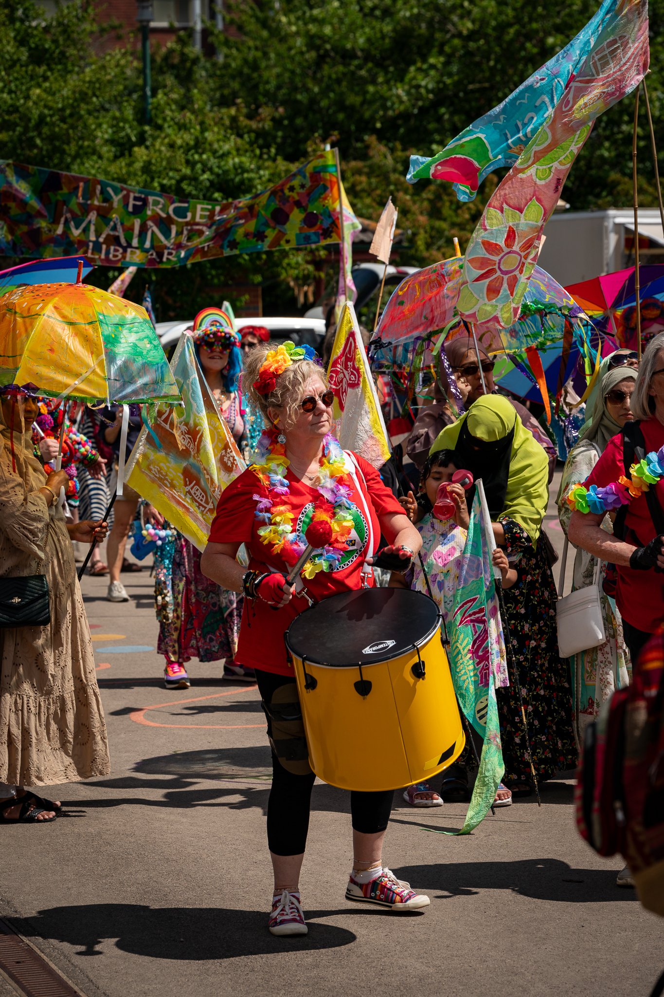 Crowd of people participating in a parade or festival, many holding colorful umbrellas and wearing vibrant costumes and accessories, including leis, flowers, and face paint, outdoors on a sunny day.