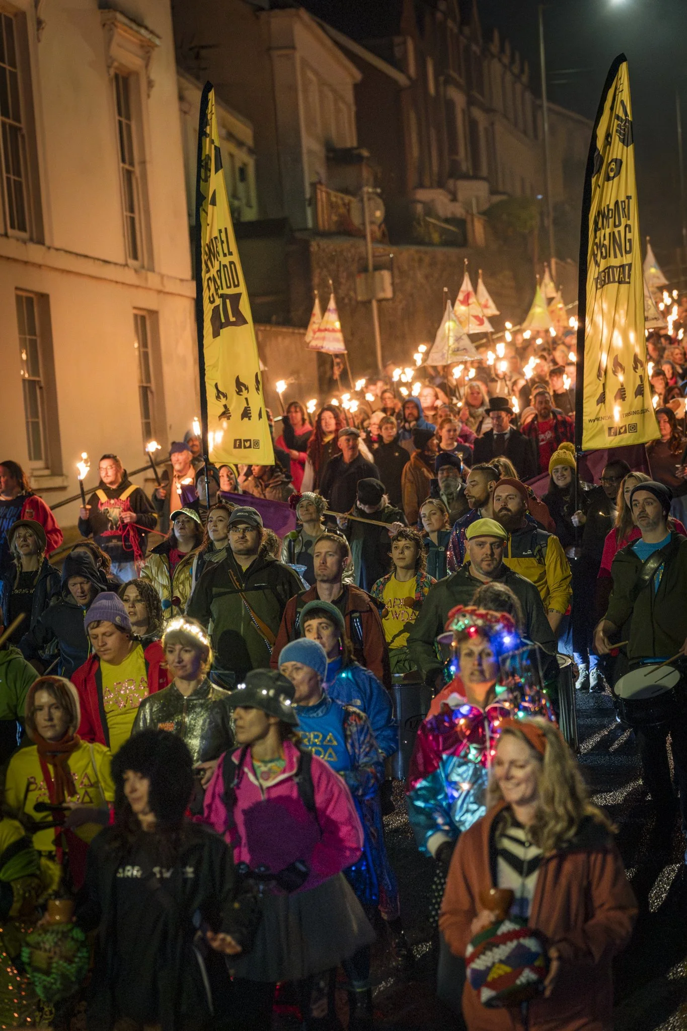 A large group of people participating in a nighttime torch-lit parade on a city street, with some holding flags and wearing colorful clothing.