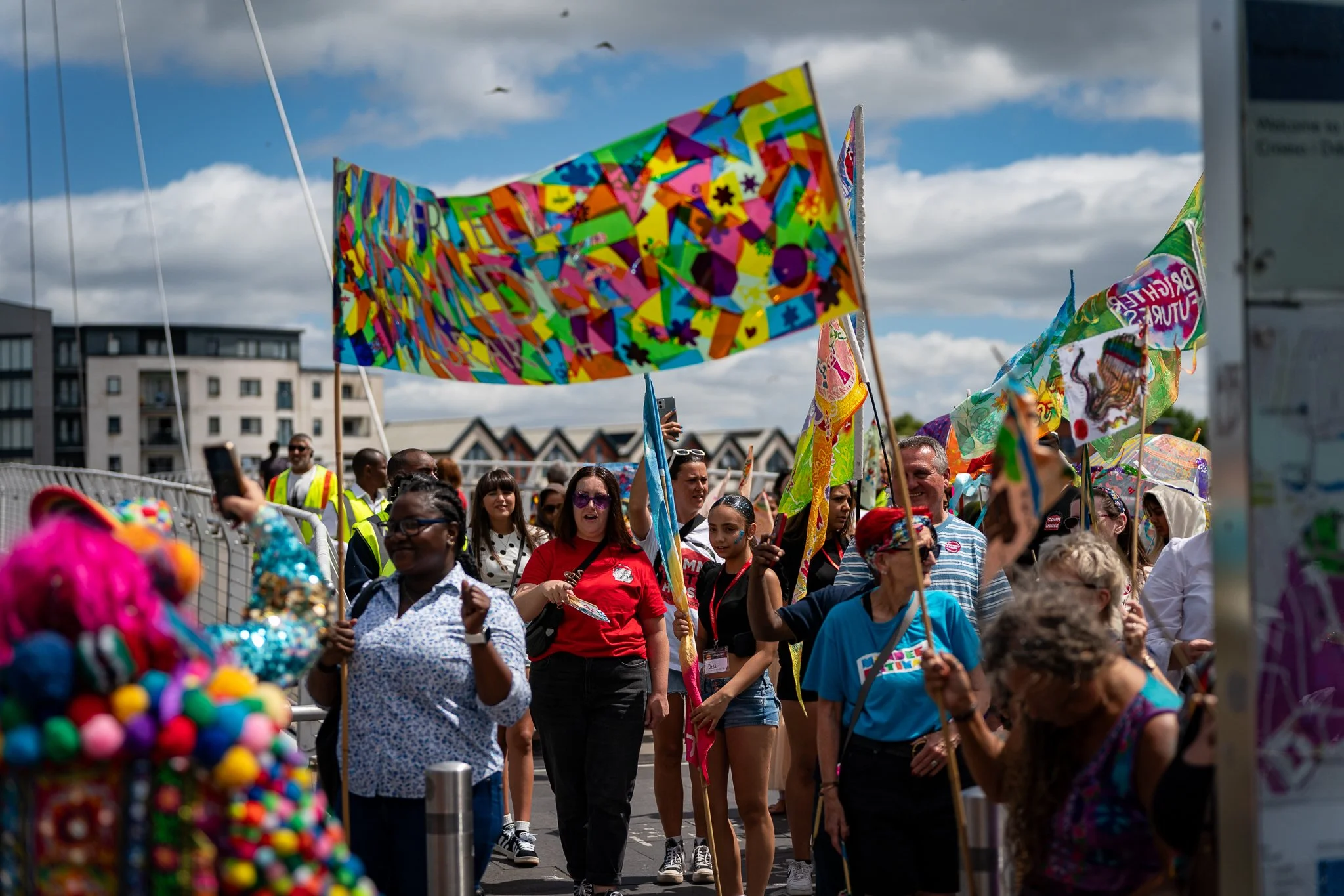 People marching in a parade, holding colorful flags and banners, with a cloudy sky and modern buildings in the background.