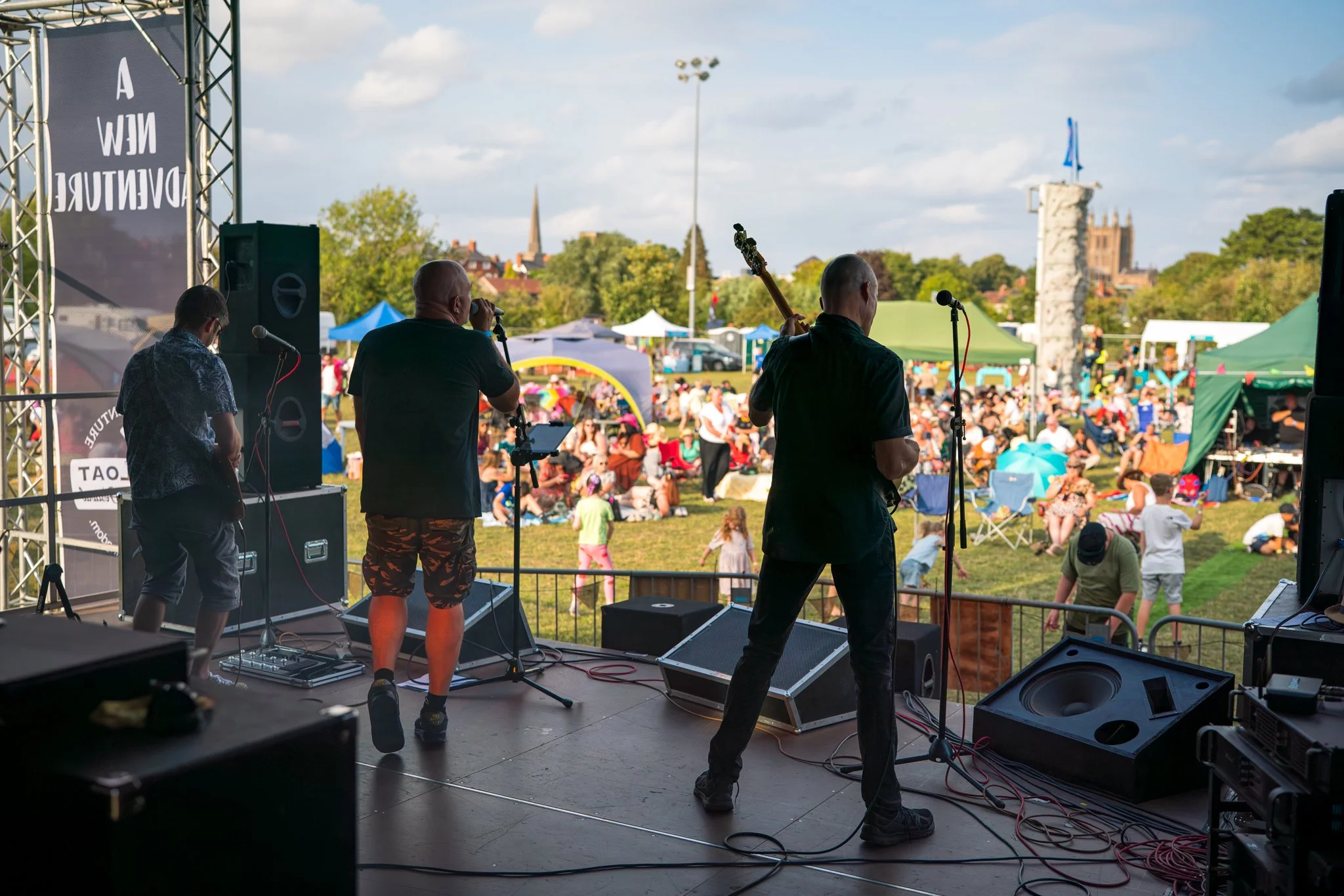 Musicians performing on an outdoor stage at a festival with a large crowd and tents in the background.