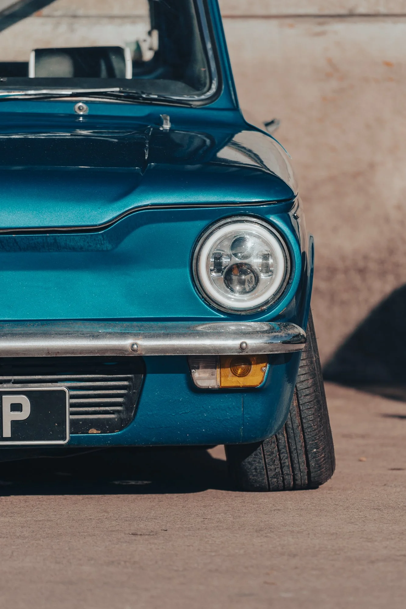 Close-up of the front of a vintage blue car parked on a dirt road, focusing on the right headlight, bumper, and part of the front tire.