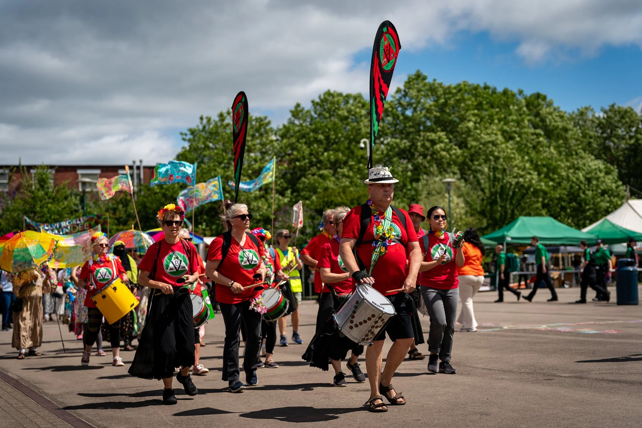 A group of people participating in a parade or festival, walking outdoors on a sunny day. Many are wearing red shirts with a green and white emblem, playing drums, and some are carrying umbrellas and flags with bright, colorful patterns.