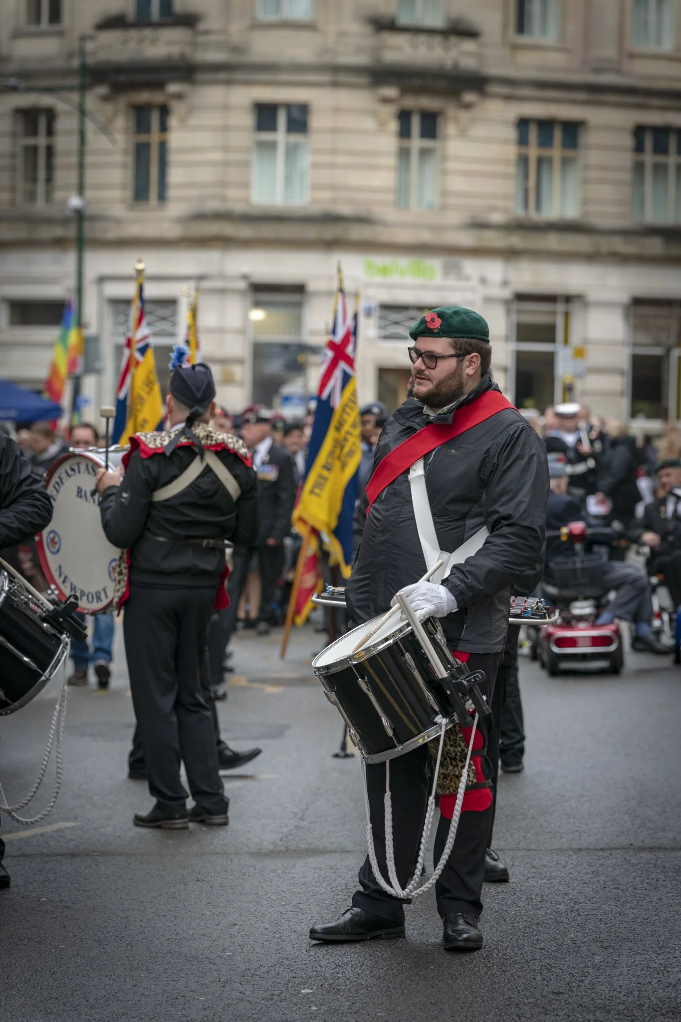 Man dressed in uniform with drum, standing in a crowd during a parade or event, wearing a green beret with a red poppy, glasses, and black jacket with red sash, holding drumsticks, among others with flags and musical instruments in an urban setting.