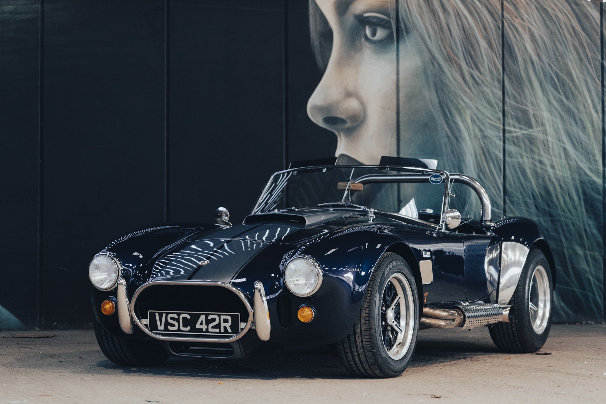 A vintage black sports car parked in front of a large mural of a woman's face with blonde hair. The car has a UK license plate reading VSC 42R and features chrome details and round headlights.