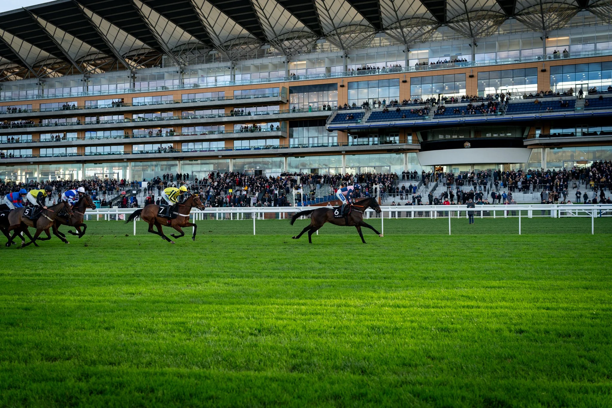 Horse race at a modern stadium with multiple levels of spectators in the stands and racehorses with jockeys in the foreground.