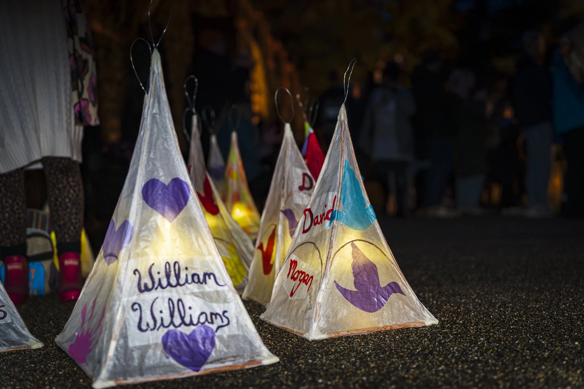 Handmade paper lanterns decorated with colorful drawings and names, displayed on a dark outdoor surface at night.