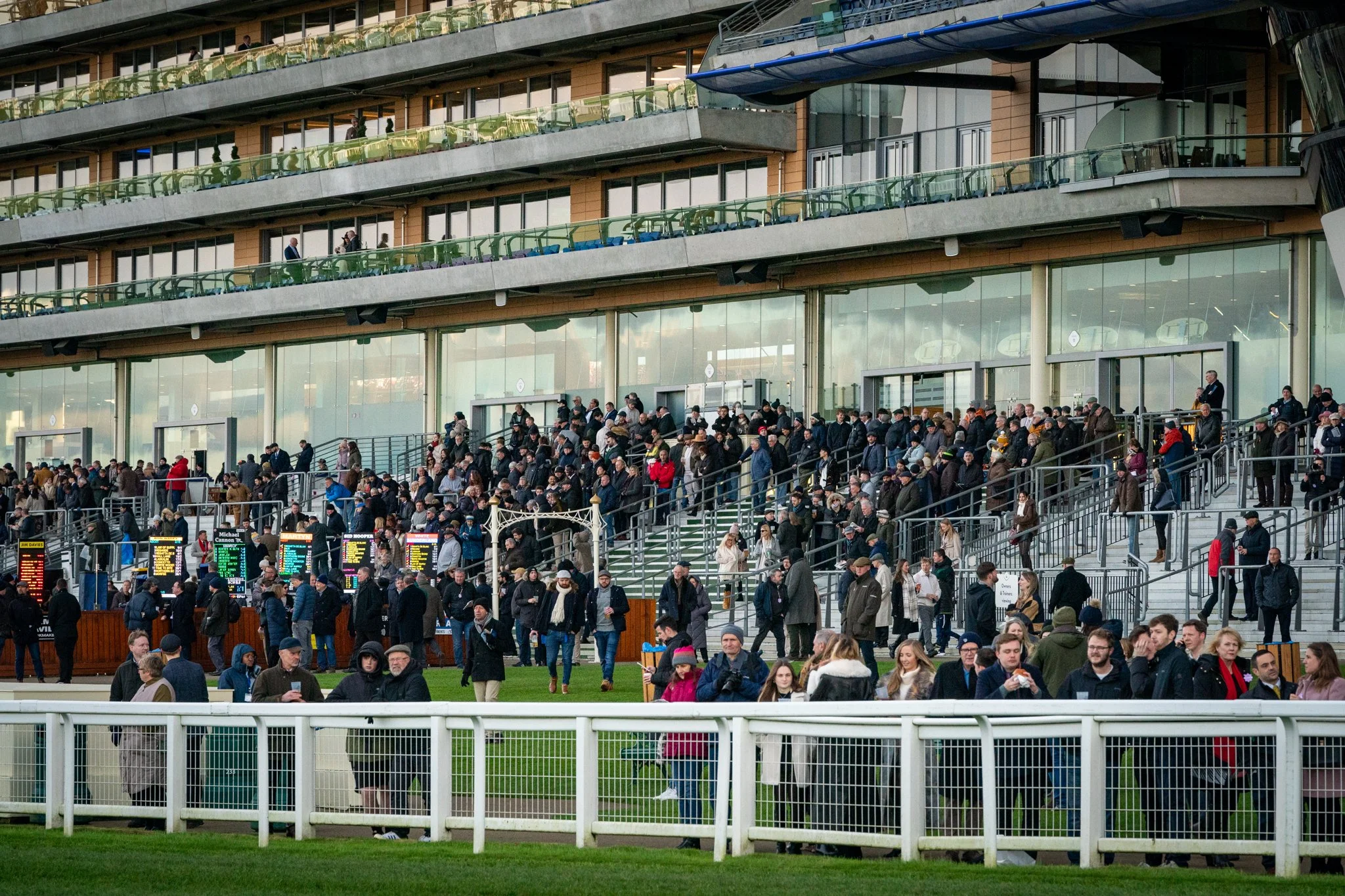 Crowd of people at a horse racing event, viewed from the stands with multiple levels of seating and some individuals walking on the ground floor outside the building.