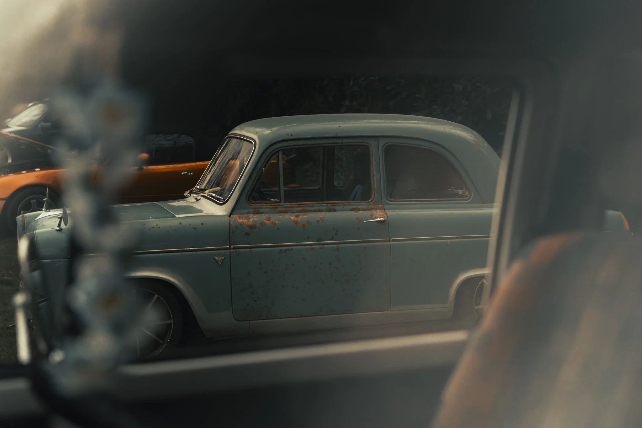 A vintage blue car with rust and wear on its body, parked among other vehicles, seen through another car's window. .