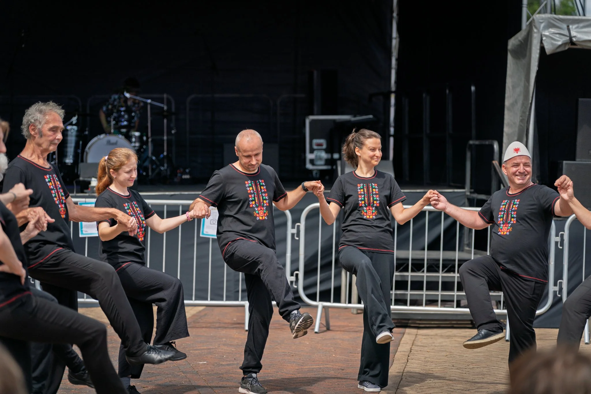 Group of people dancing in a circle on an outdoor stage, holding hands and lifting their legs in a traditional dance, wearing black shirts with colorful embroidery designed on the front.