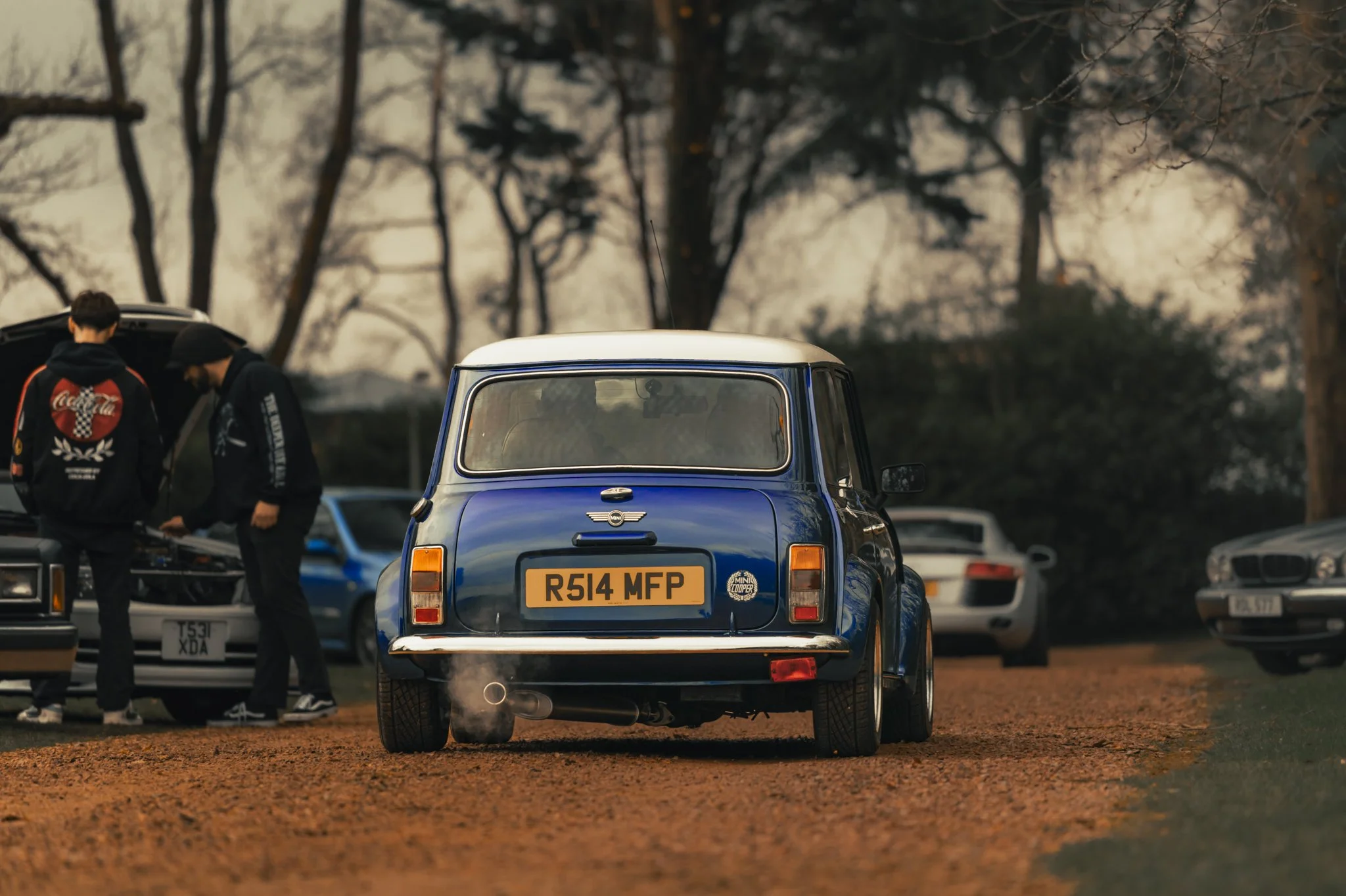 A blue vintage MINI Cooper driving on a dirt surface, with two people standing beside an open car trunk and several other cars parked in the background. The scene appears to be in an outdoor setting with leafless trees and a hazy sky.