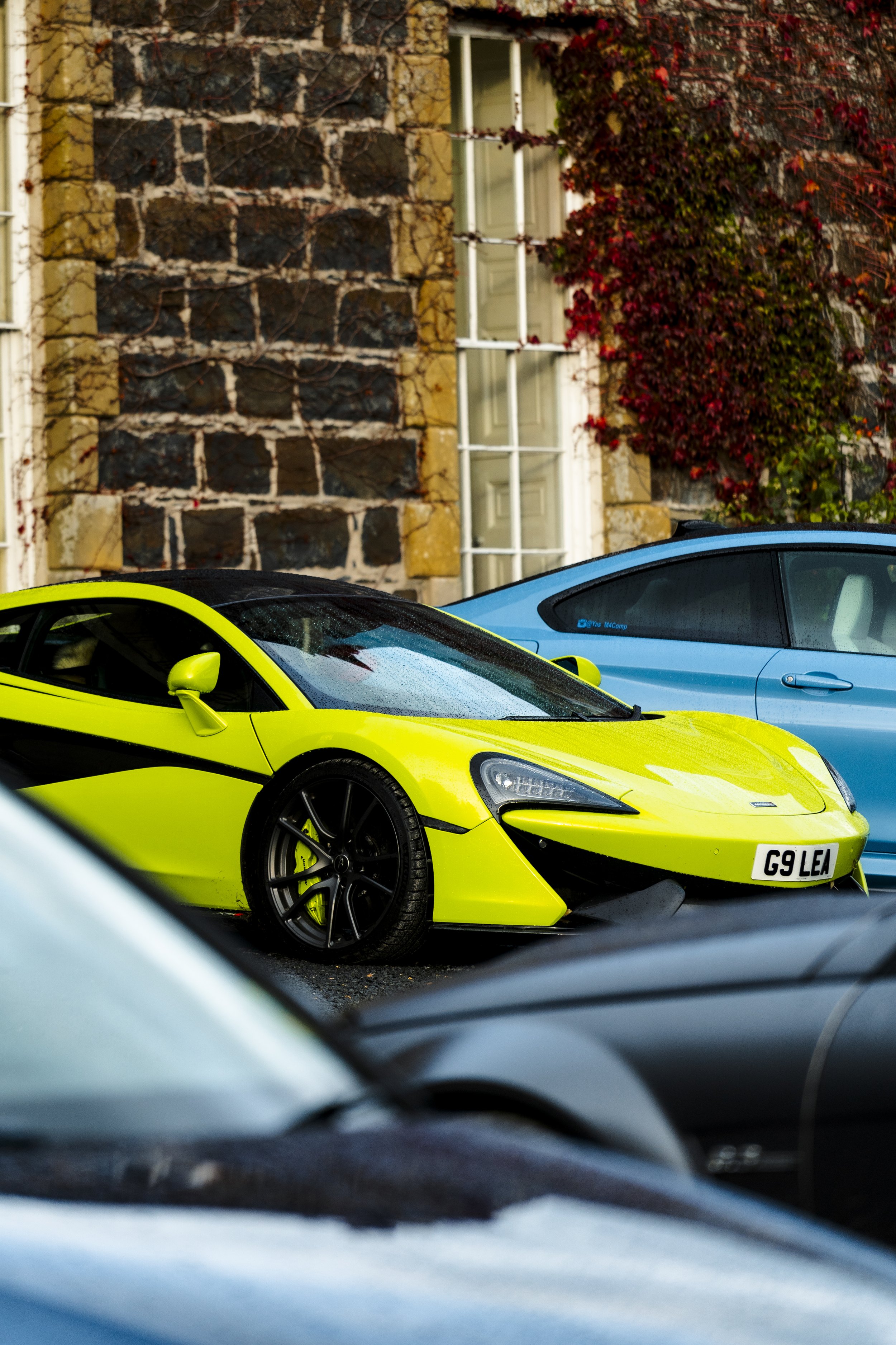 A bright yellow sports car with black accents and a UK license plate G9 LE parked on a street, with a blue sports car partially visible behind it. The background features a stone building with a window and red ivy growing on the wall.