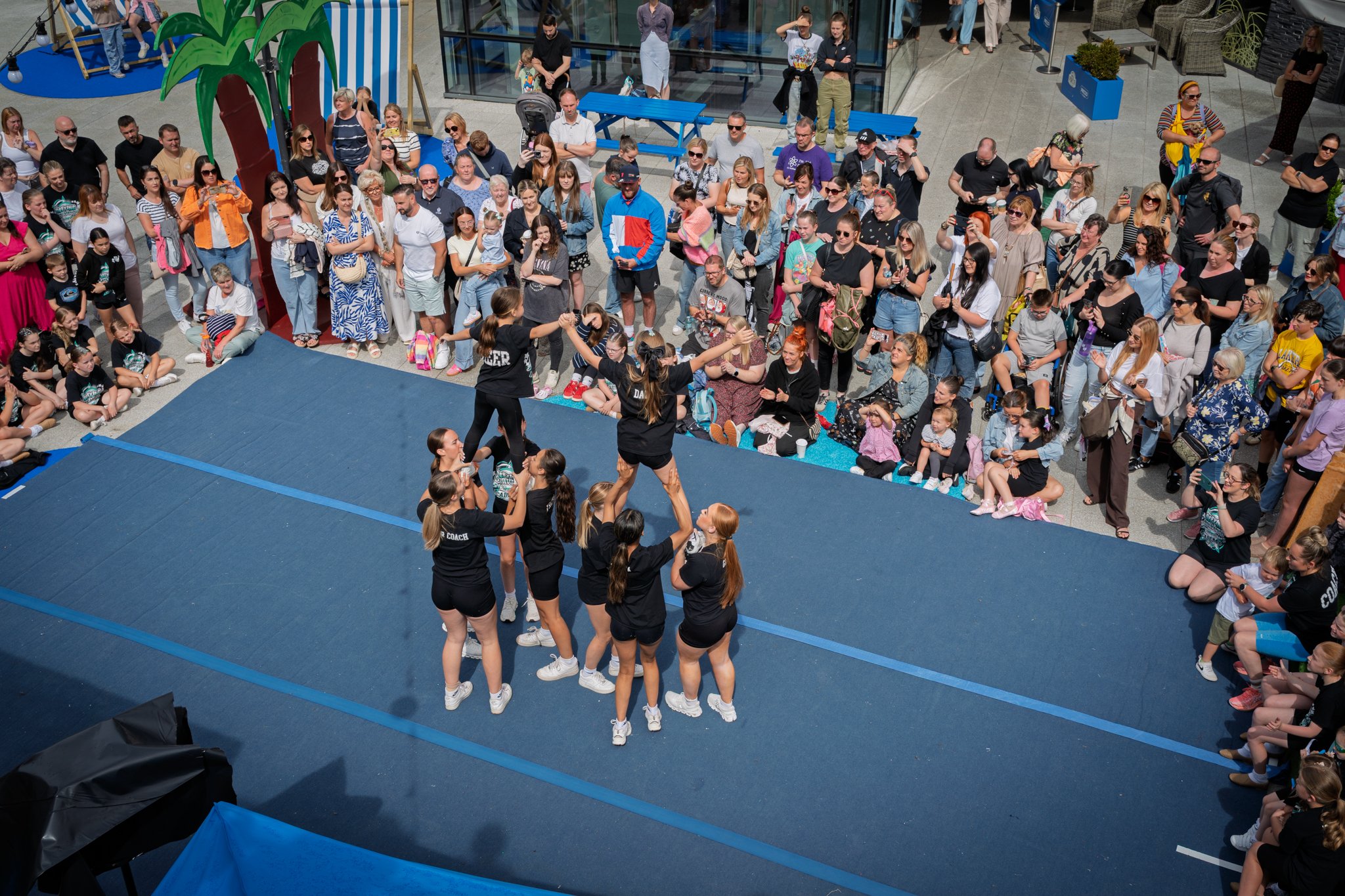 A group of cheerleaders or dancers performing on a blue mat in front of a large crowd at an indoor or outdoor event, with spectators standing and sitting around the performance area.