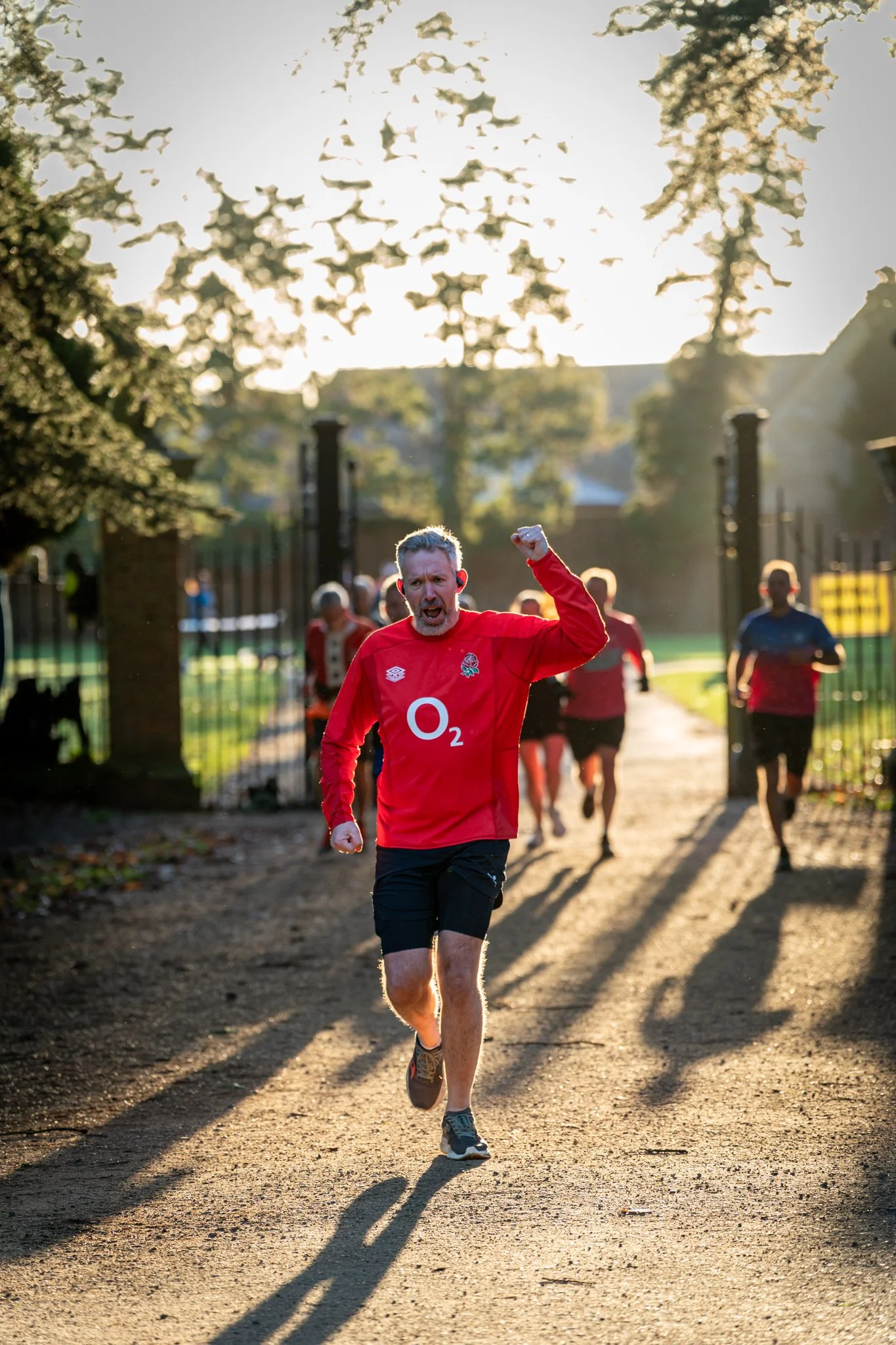 A man in a red sports jersey and black shorts is running on a dirt path outdoors, raising his right fist in celebration. Several other runners are visible behind him, and the scene is backlit by sunlight, casting long shadows and creating a warm glow