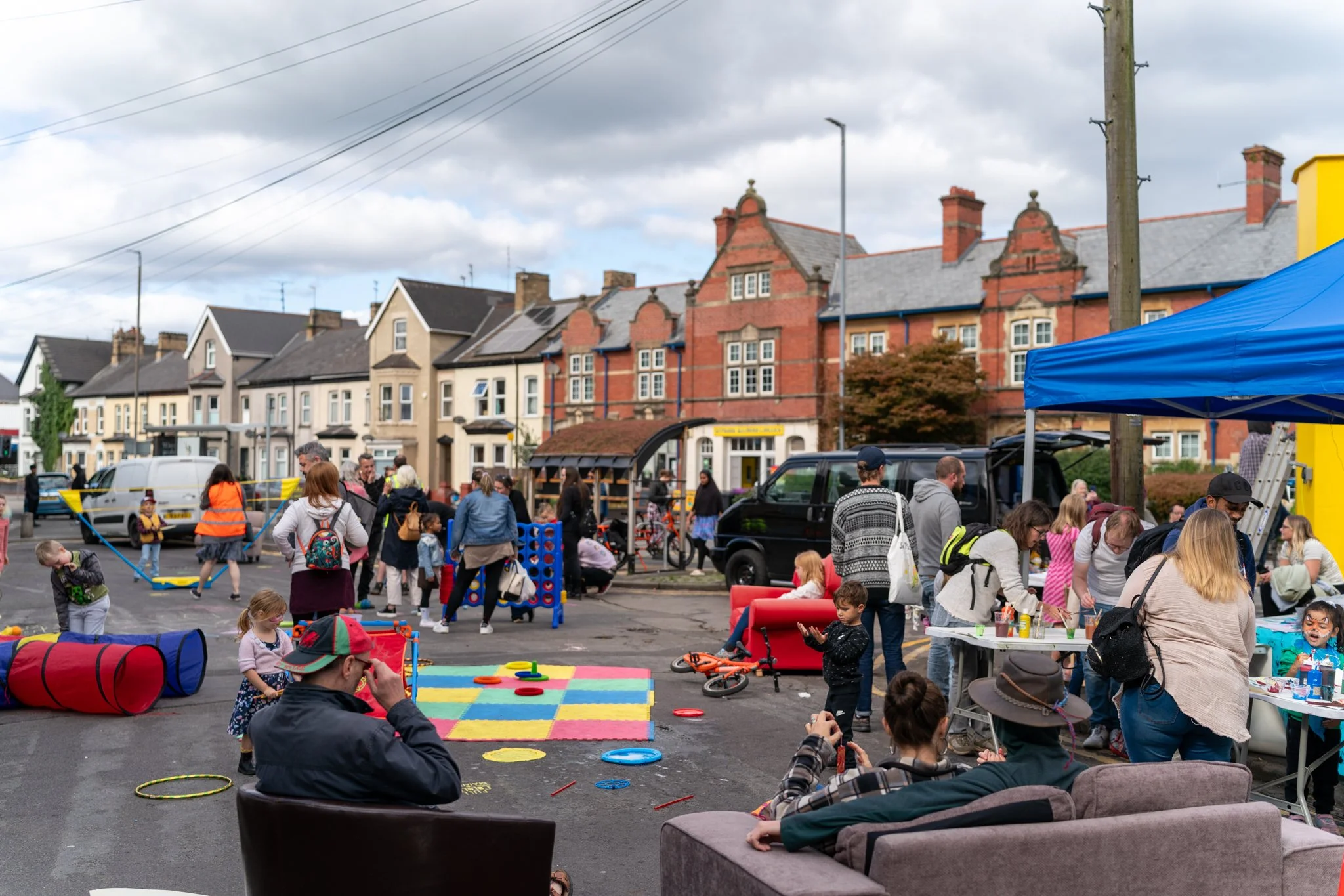 A community gathering in a town square with children playing, adults socializing, and various activities. There are colorful toys, a large rainbow mat, and tents set up on the street, with row houses and cloudy sky in the background.