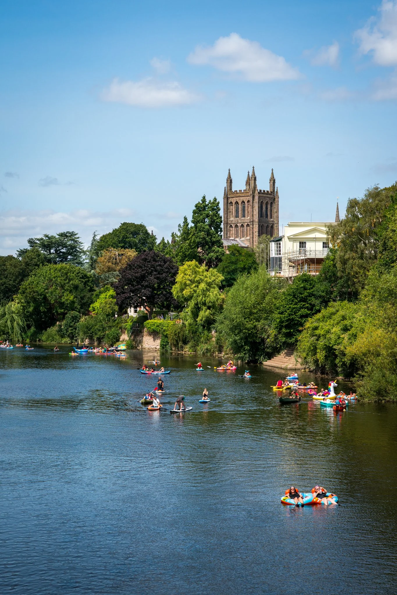 People floating and paddling on the river with a historic church and trees in the background on a sunny day.