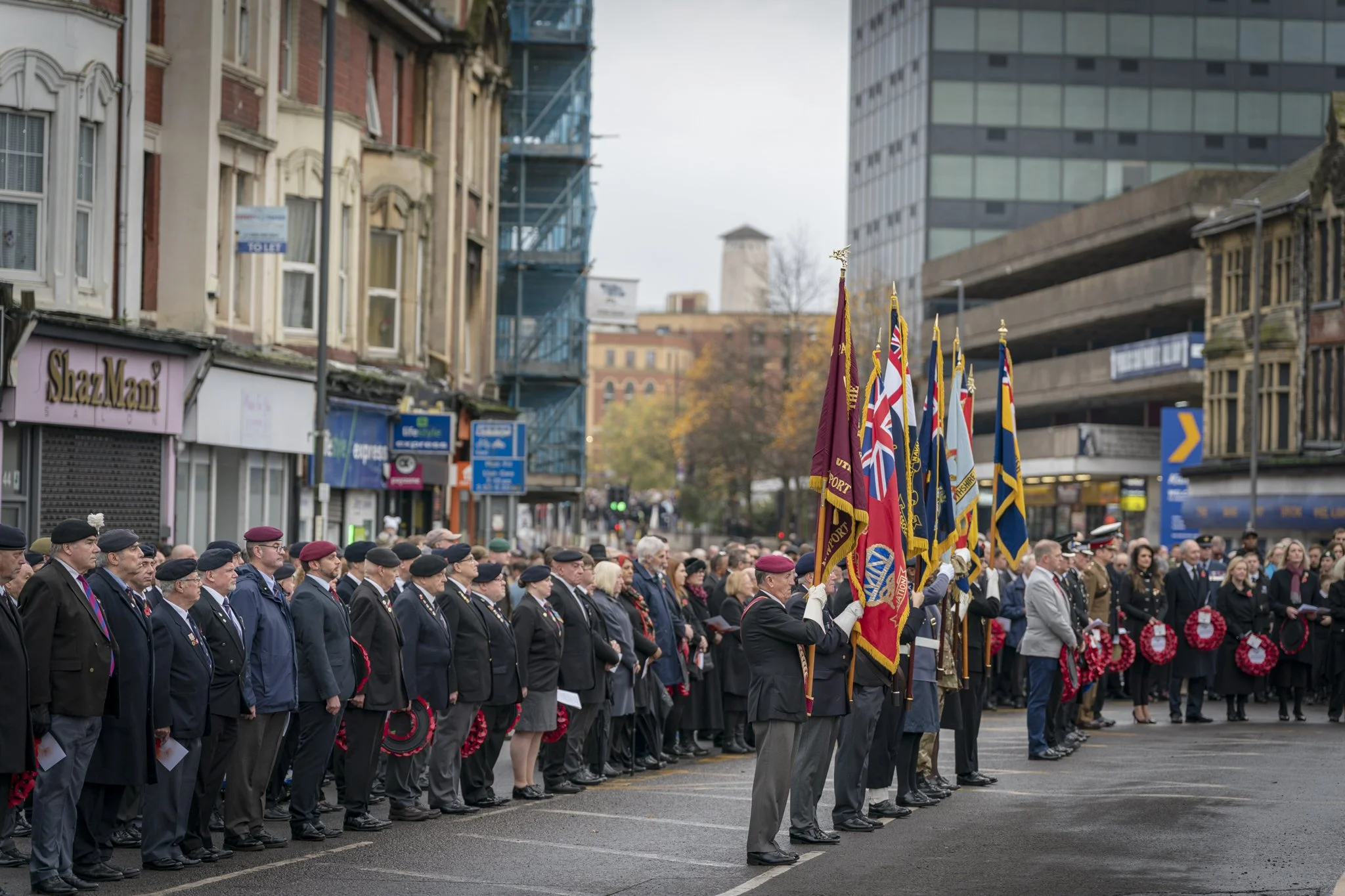 A large group of people dressed in formal attire, standing in formation during a memorial or commemorative event on a city street, with flags held by some participants. The background shows urban buildings, some under construction and others older.
