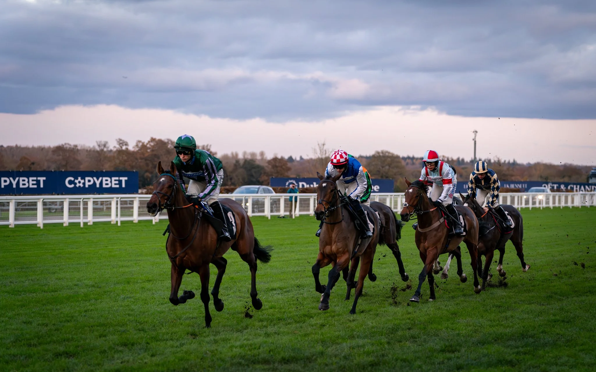Horse race with jockeys in colorful uniforms on a grassy racecourse under cloudy skies.