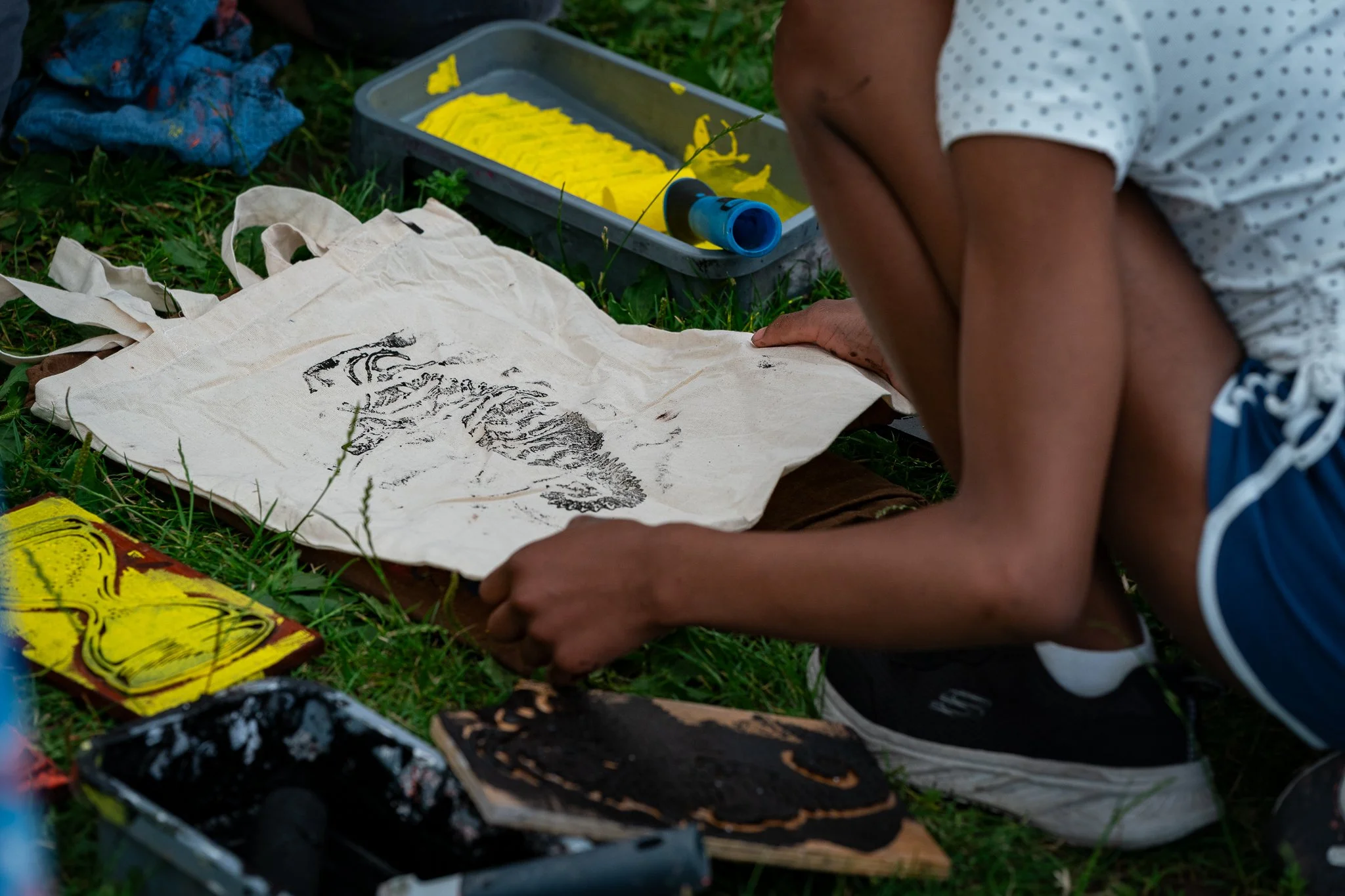 Person creating a fossil rubbing of a dinosaur skeleton on a piece of paper outdoors, surrounded by art supplies and grass.