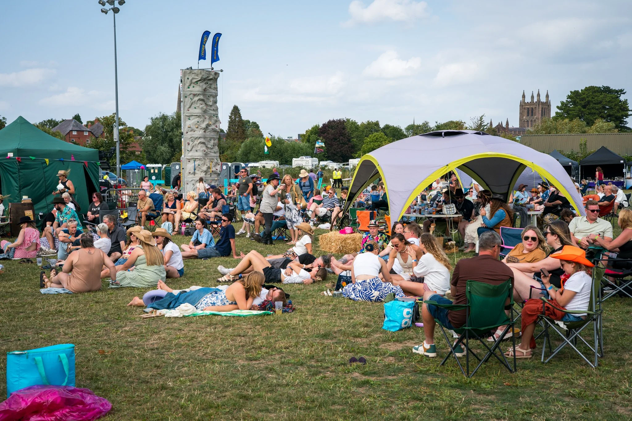People gathered outdoors at a festival or event with tents, a climbing wall, and a stage, on a sunny day with blue skies and a church in the background.