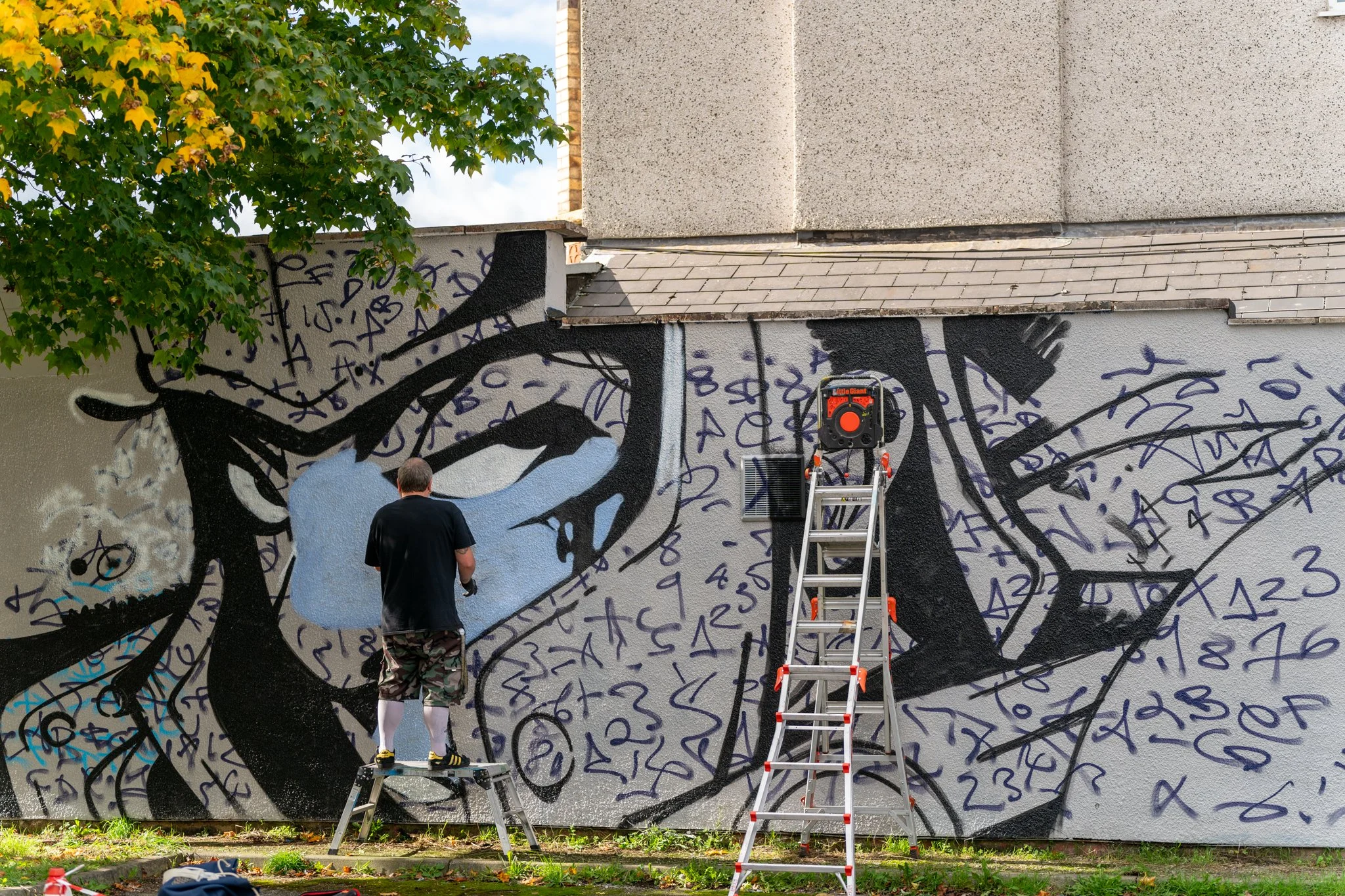 A person standing in front of a colorful mural with black, white, and purple abstract design and numbers. Two ladders are positioned near the mural, with one person on one ladder and another standing nearby. Green trees and a building are in the back