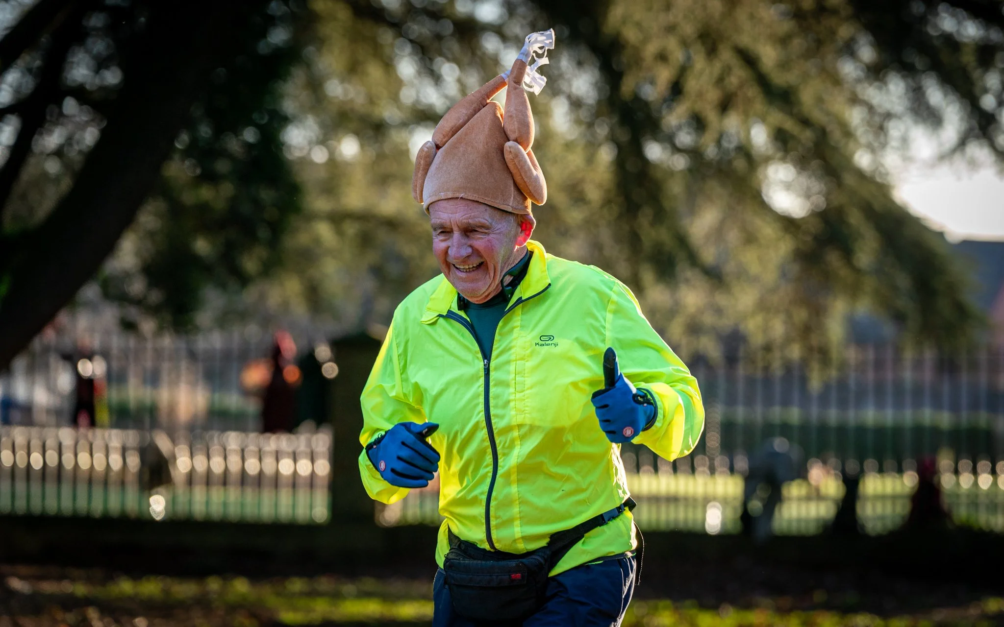 An elderly man running outdoors during daylight, wearing a bright yellow jacket, blue gloves, and a beige hat with a decorative puffed design. He appears to be smiling or enjoying his run.