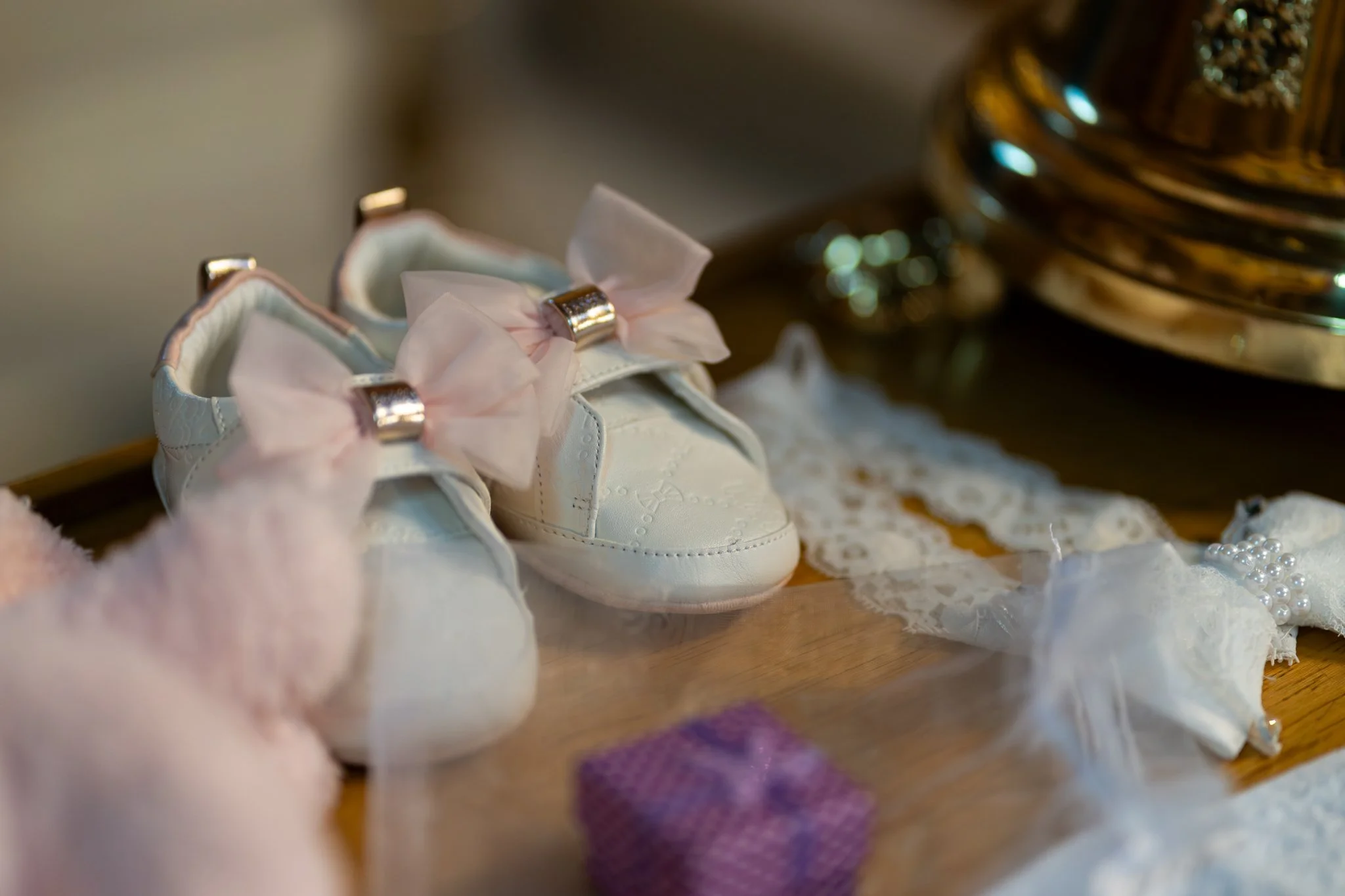 Close-up of white infant shoes decorated with pink bows and silver accents, placed on a lace cloth with various small accessories and fabric items nearby.