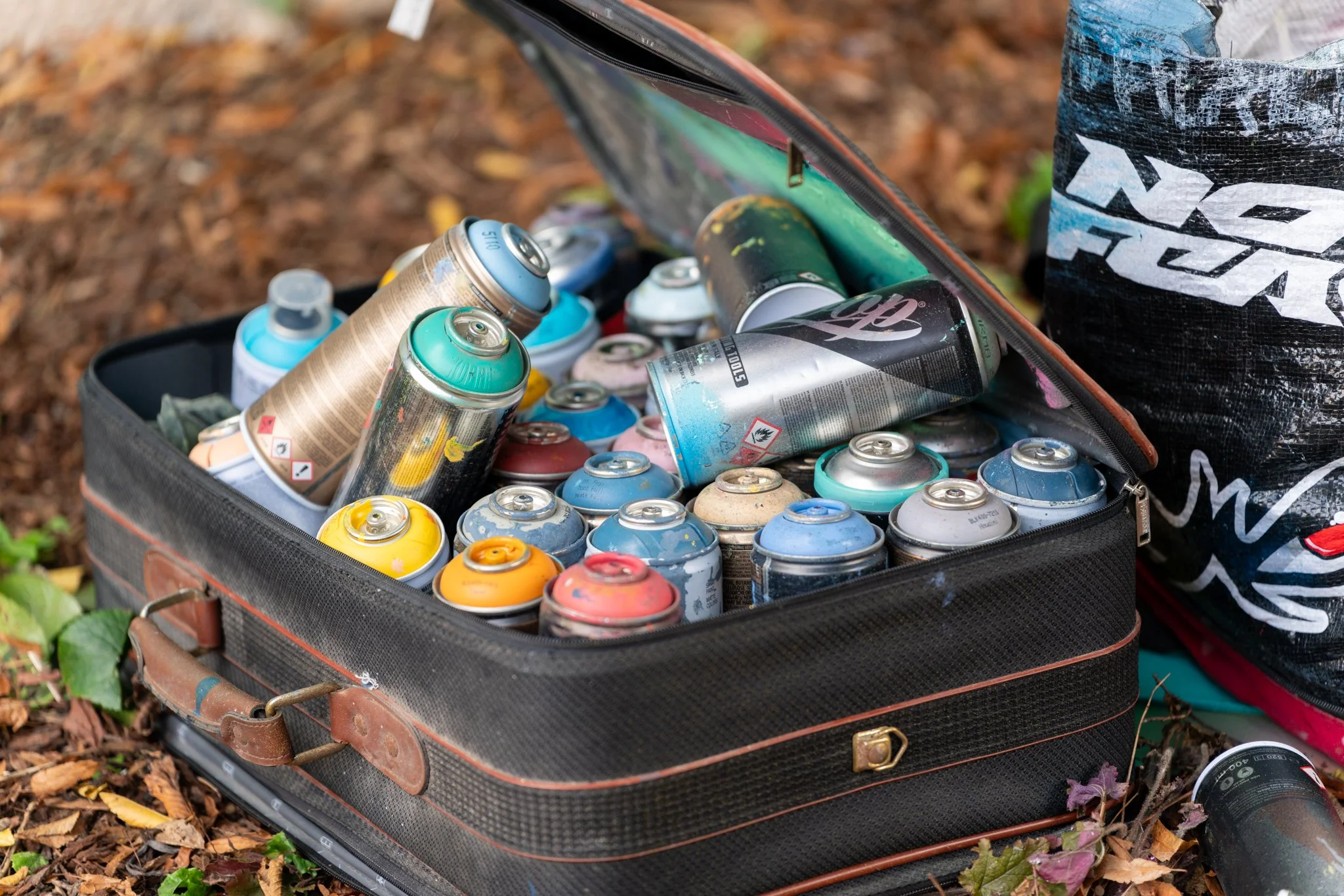 A vintage suitcase filled with spray paint cans, open on autumn leaves and grass, with a black and white plastic bag nearby.