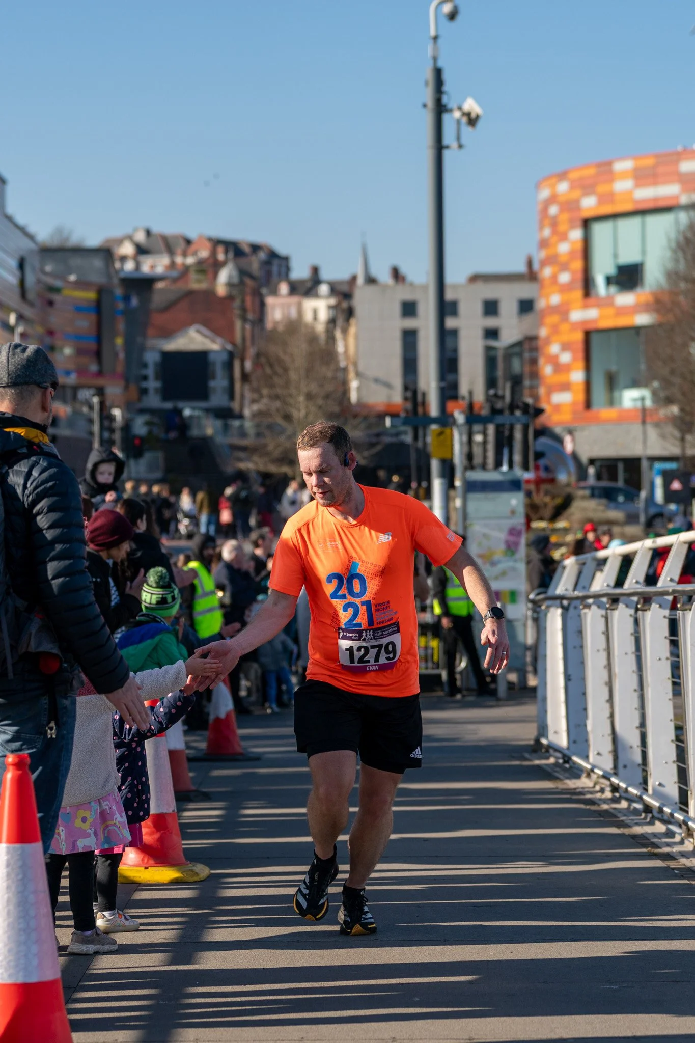 A male runner in an orange shirt and black shorts crossing a finish line, shaking hands with a young girl, with spectators and a cityscape in the background.