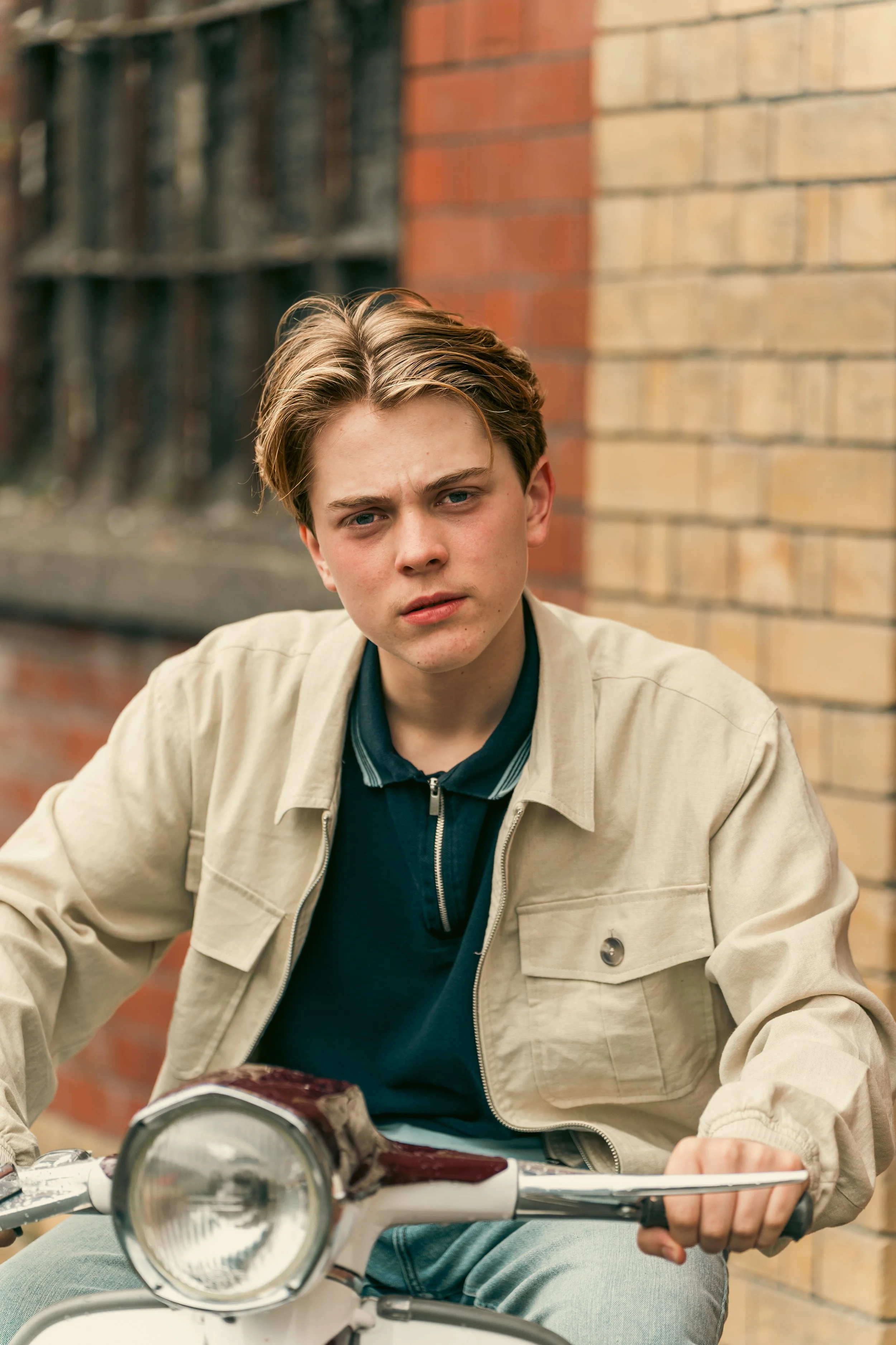 Young man sitting on a motorcycle, looking at the camera, against a background of a brick wall.