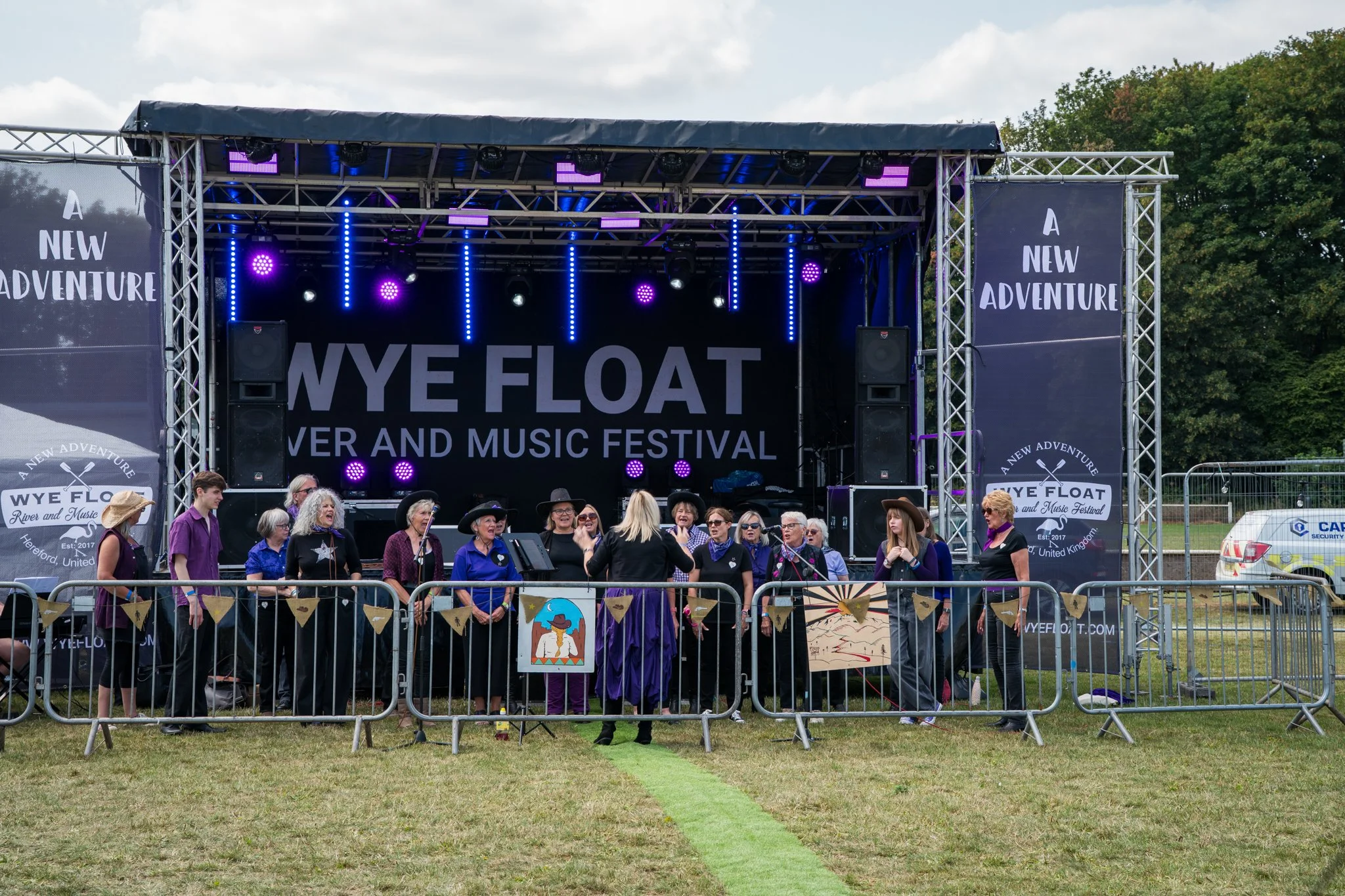 A group of people singing on a stage at an outdoor festival with a large sign reading 'NYE FLOAT Summer and Music Festival' and banners that say 'A New Adventure' and 'WYEFLOAT' in the background.