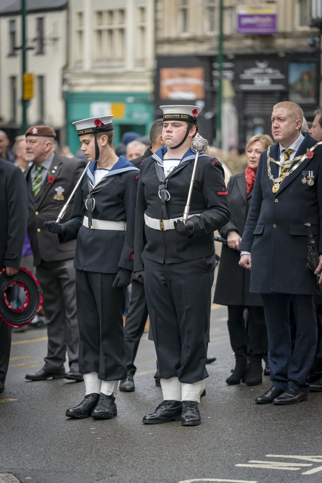 Two young sailors in navy uniforms marching in a parade, accompanied by adults in formal attire, on a city street with onlookers and buildings in the background.