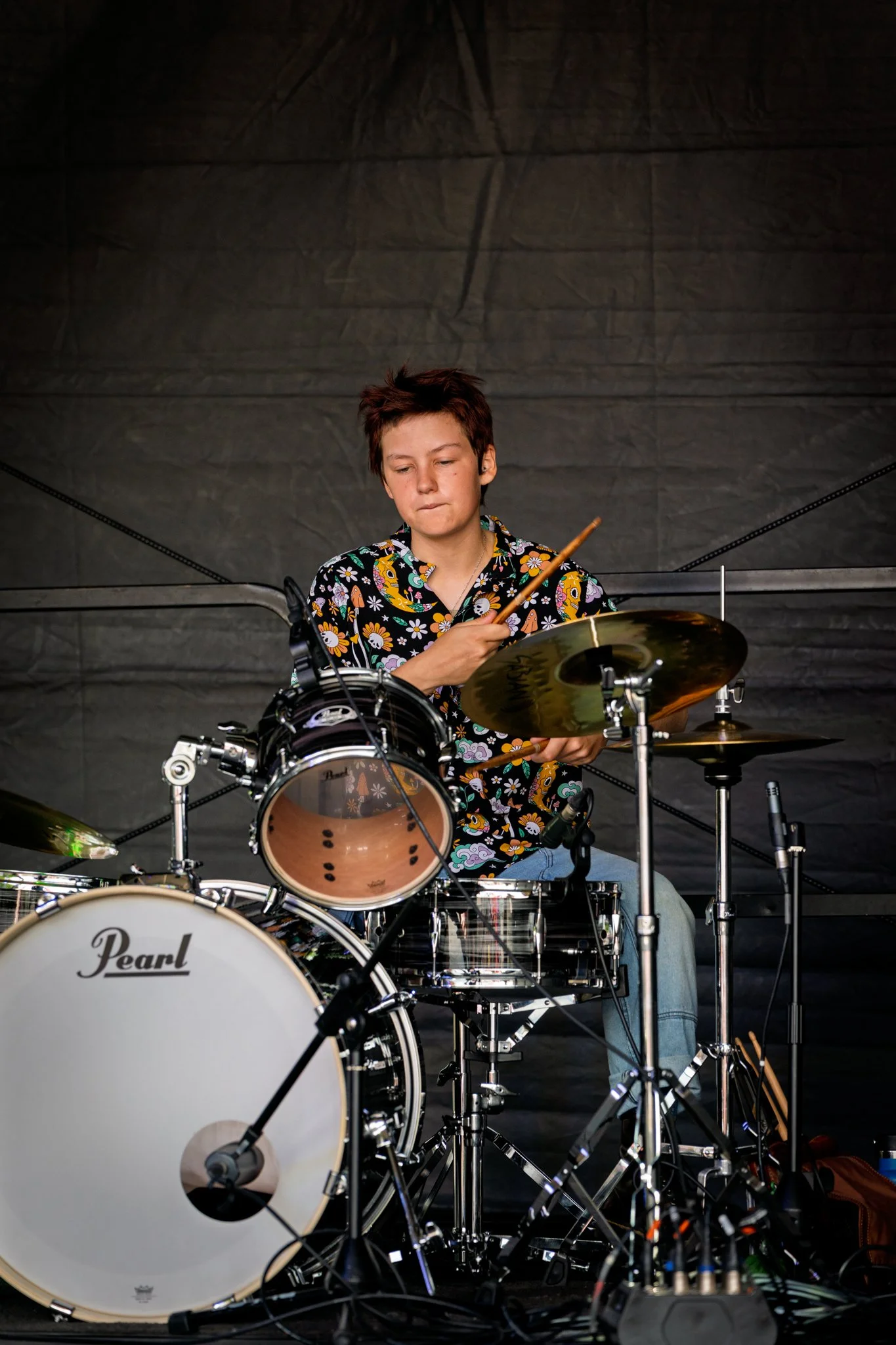 A person with short dark hair playing a drum set, wearing a black floral shirt, against a dark background.