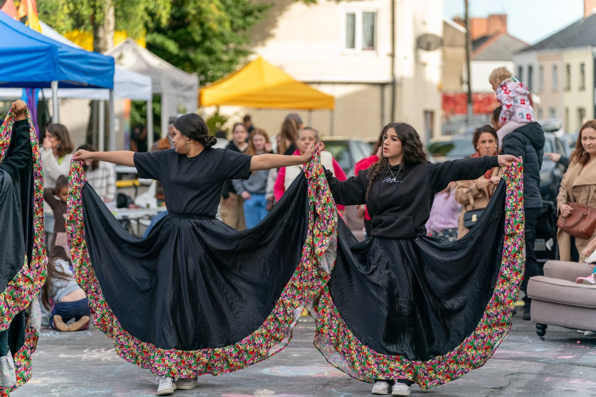 Two women dancing outdoors under tents, holding floral skirts as they twirl, with onlookers in the background.