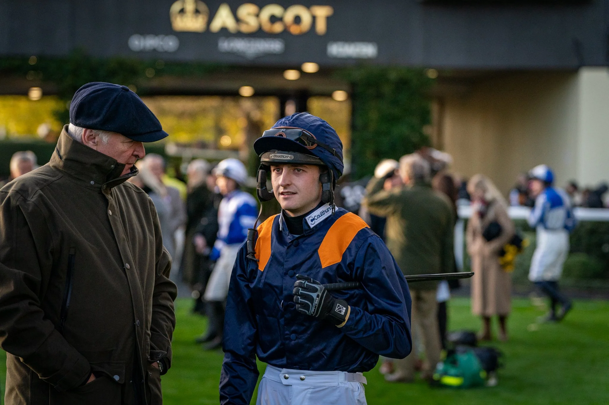 A man wearing a brown jacket and a black cap standing next to a young jockey dressed in blue racing attire, including a helmet, at a racetrack with a crowd in the background.