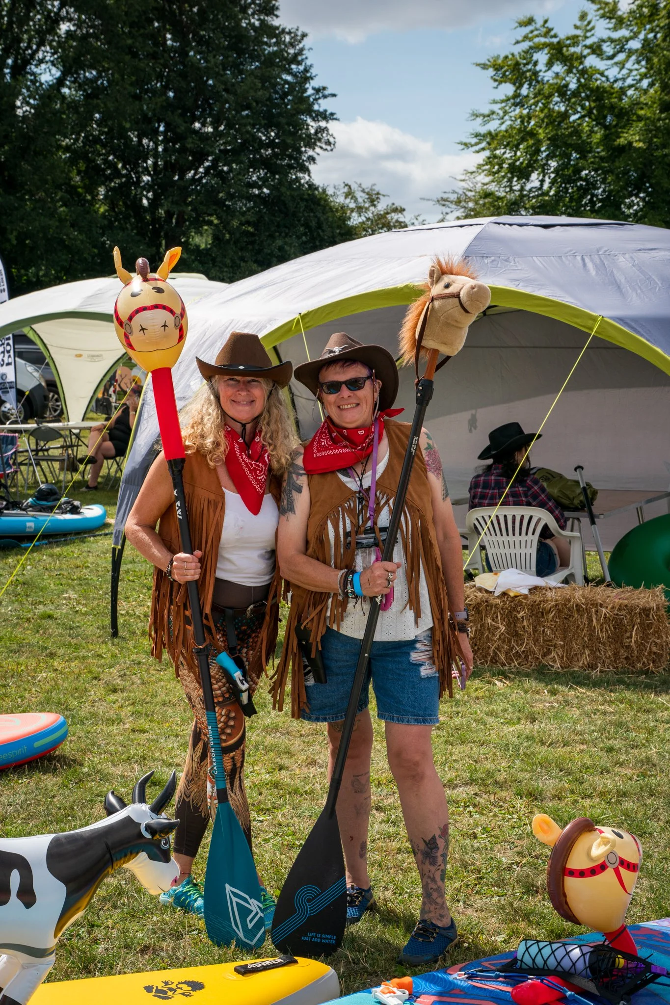 Two women dressed as cowboys standing outdoors at a festival or event, holding toy lances with horse and animal figures, smiling, with tents, trees, and other festival attendees in the background.