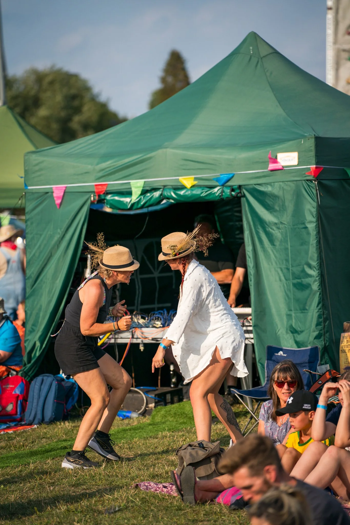People gathered outdoors at an event with a green tent overhead, some are sitting on the grass and others are dancing or socializing.