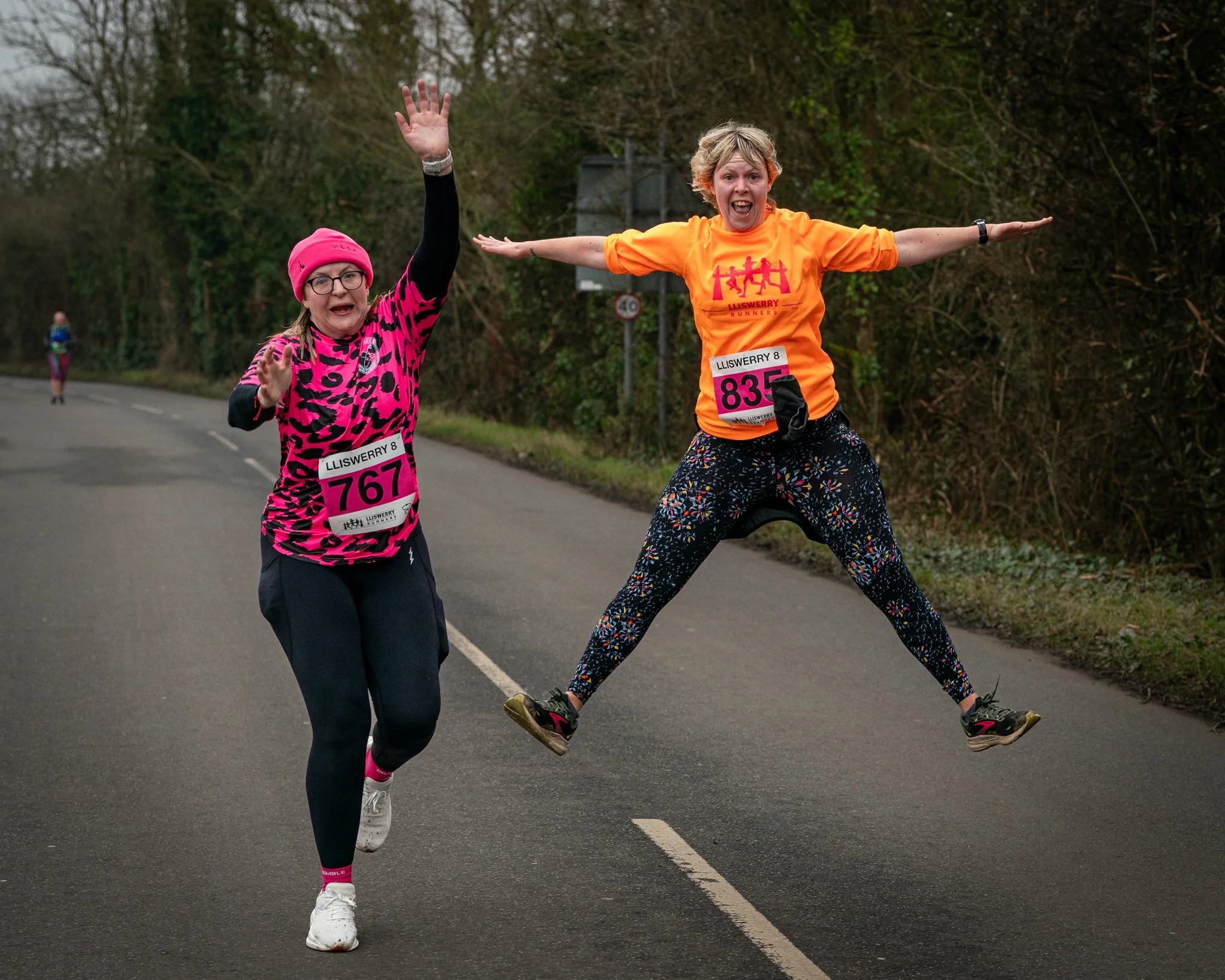Two women wearing colorful running shirts and bib numbers participating in a race on a rural road surrounded by trees, with one woman jumping with arms outstretched and the other waving.