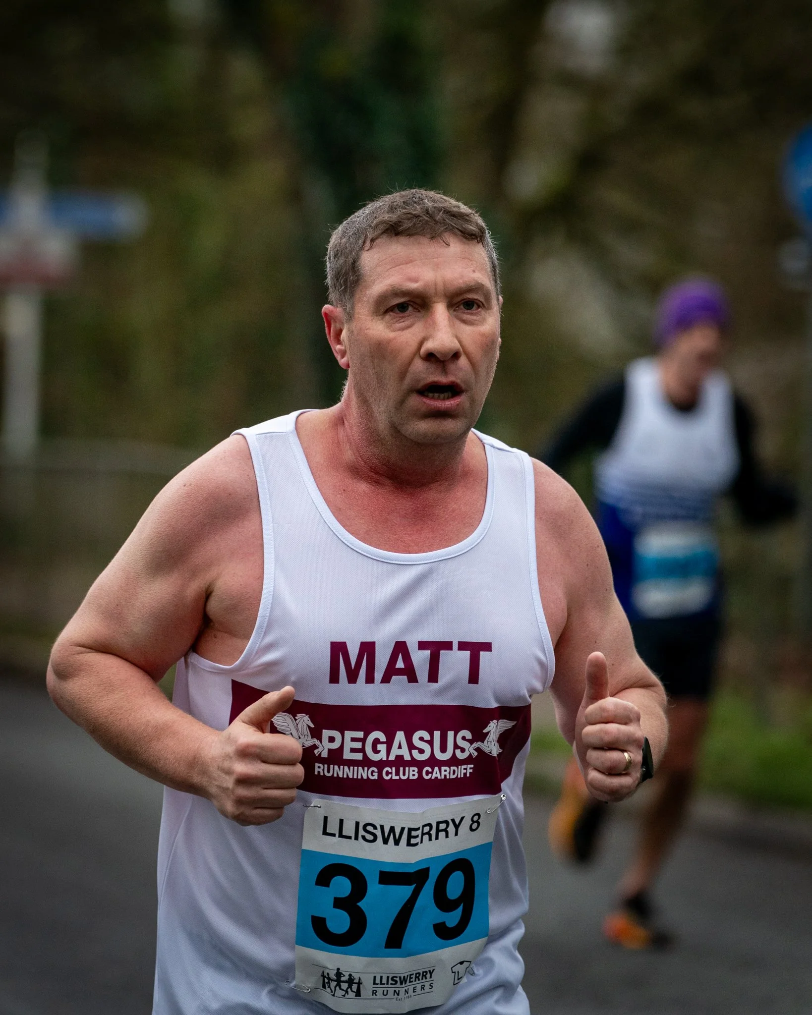 Man running in a race wearing a white tank top with a bib number 379, with a logo and the words 'Pegasus Running Club Cardiff' and 'Lliswerry 8.' In the background, a woman with purple hair, black shirt, and white vest is also running, both on a pave