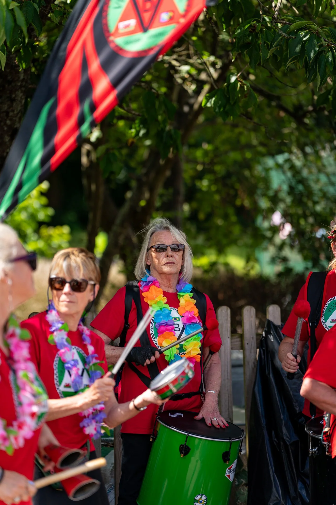 Group of older women wearing red shirts and colorful lei necklaces, holding musical instruments, outdoors with green trees in the background.