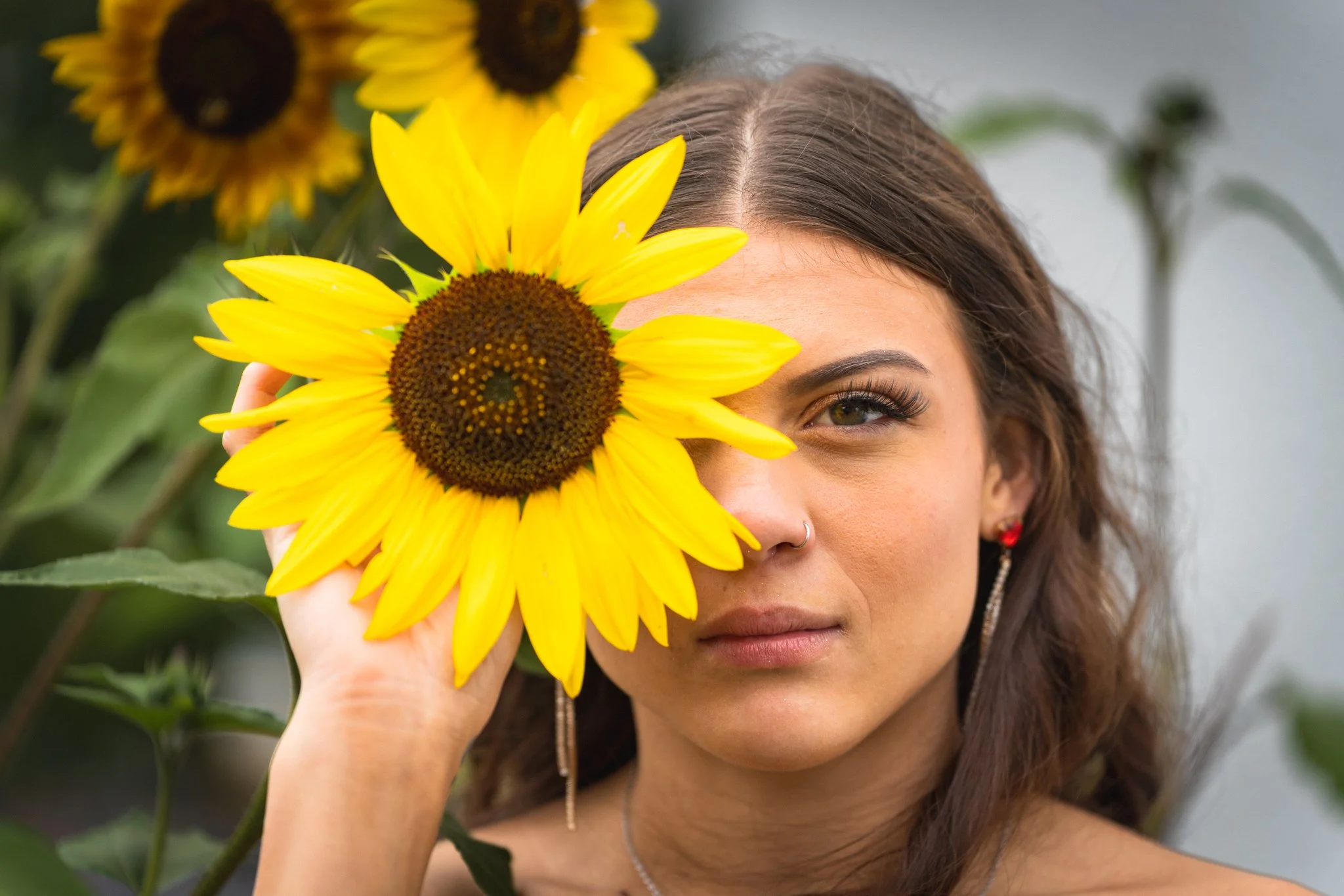 A woman holding a sunflower near her face, covering one eye, with a blurred background of sunflower plants.