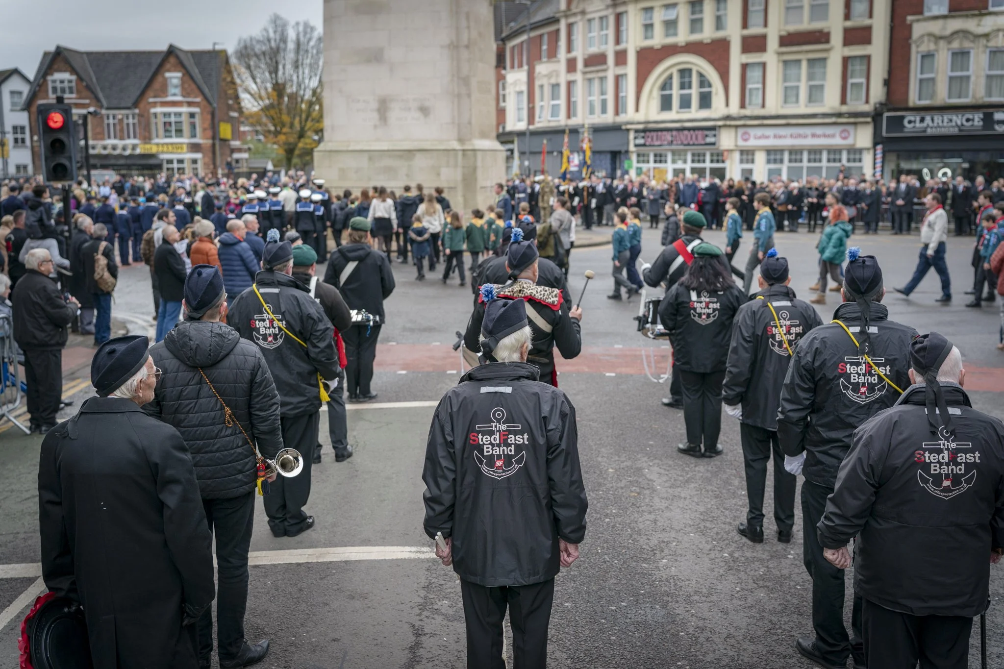 A crowd of people gathered in a city square for a parade or event, with some wearing uniforms and others casually dressed. A band with jackets labeled 'The Stedfast Band' is visible in the foreground, with some members holding instruments. A monument